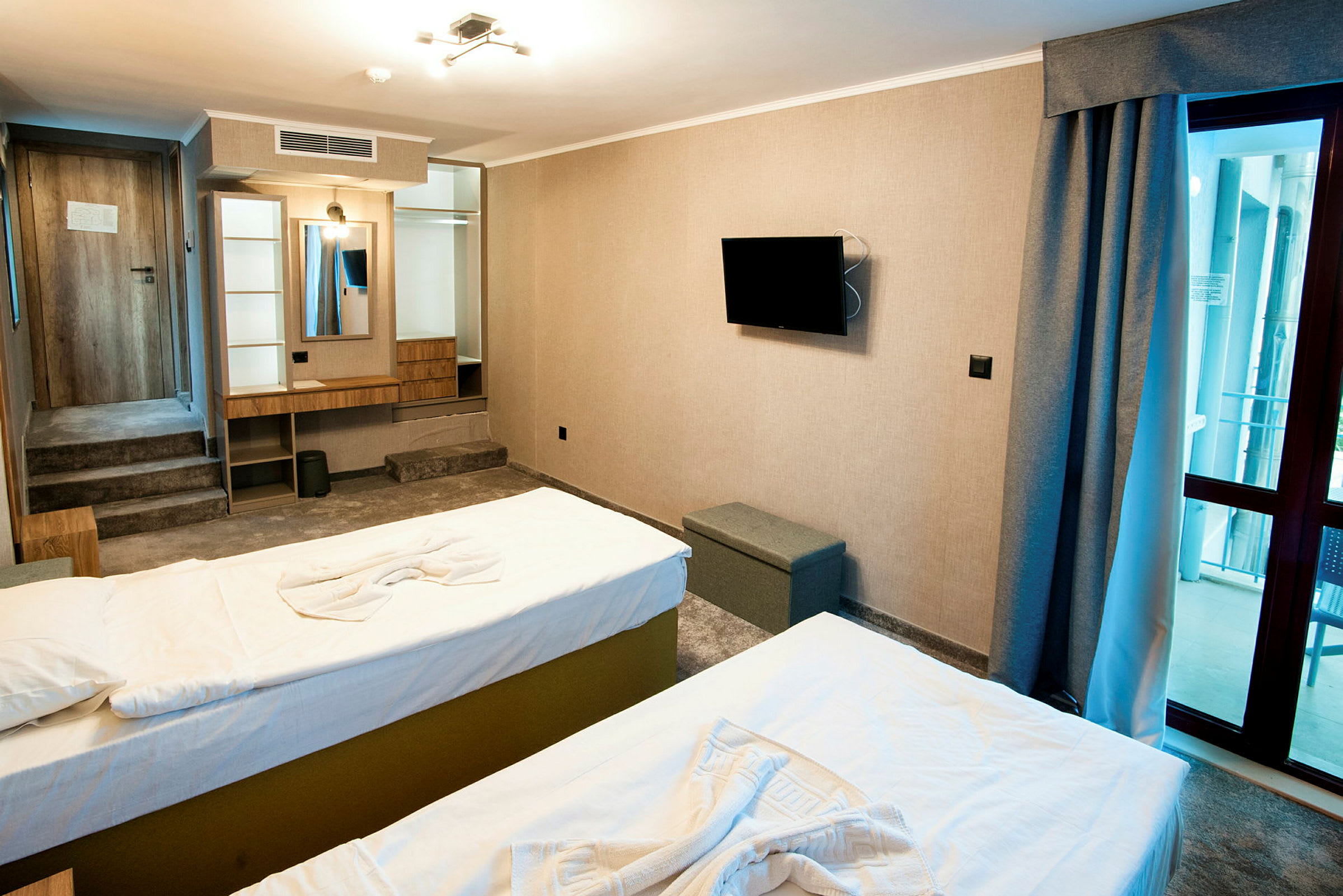 Hotellihuone, jossa on kaksi sänkyä, seinälle asennettu tv, peili ja kirjoituspöytä sekä lasiovi parvekkeelle. (Tekoälyn luoma teksti)