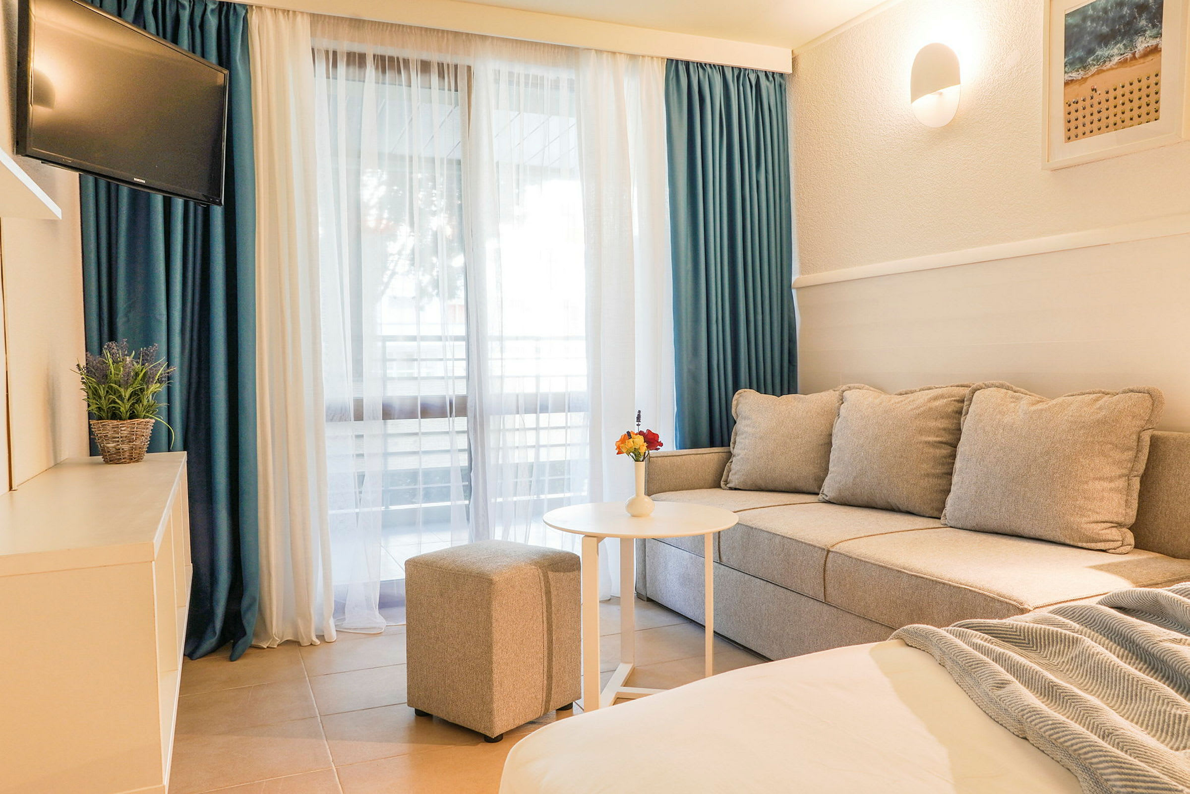 Hotellihuone, jossa on sohva, pöytä, rahi ja televisio sekä suuret ikkunat verhoineen. (Tekoälyn luoma teksti)