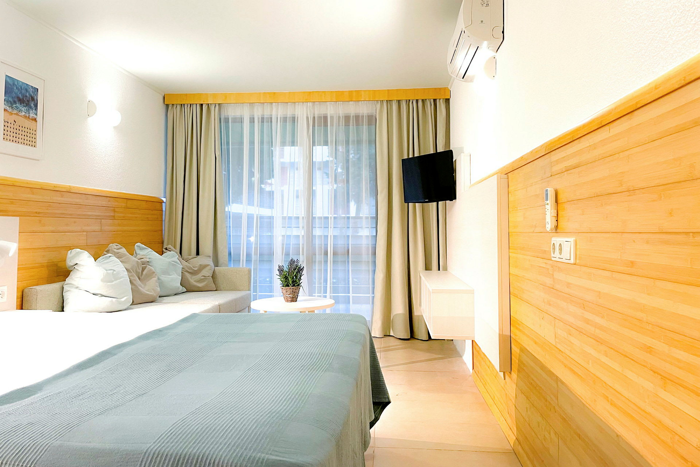 Hotellihuone, jossa on sänky, sohva ja pöytä. Verhot roikkuvat suuren ikkuna-alueen edessä. (Tekoälyn luoma teksti)