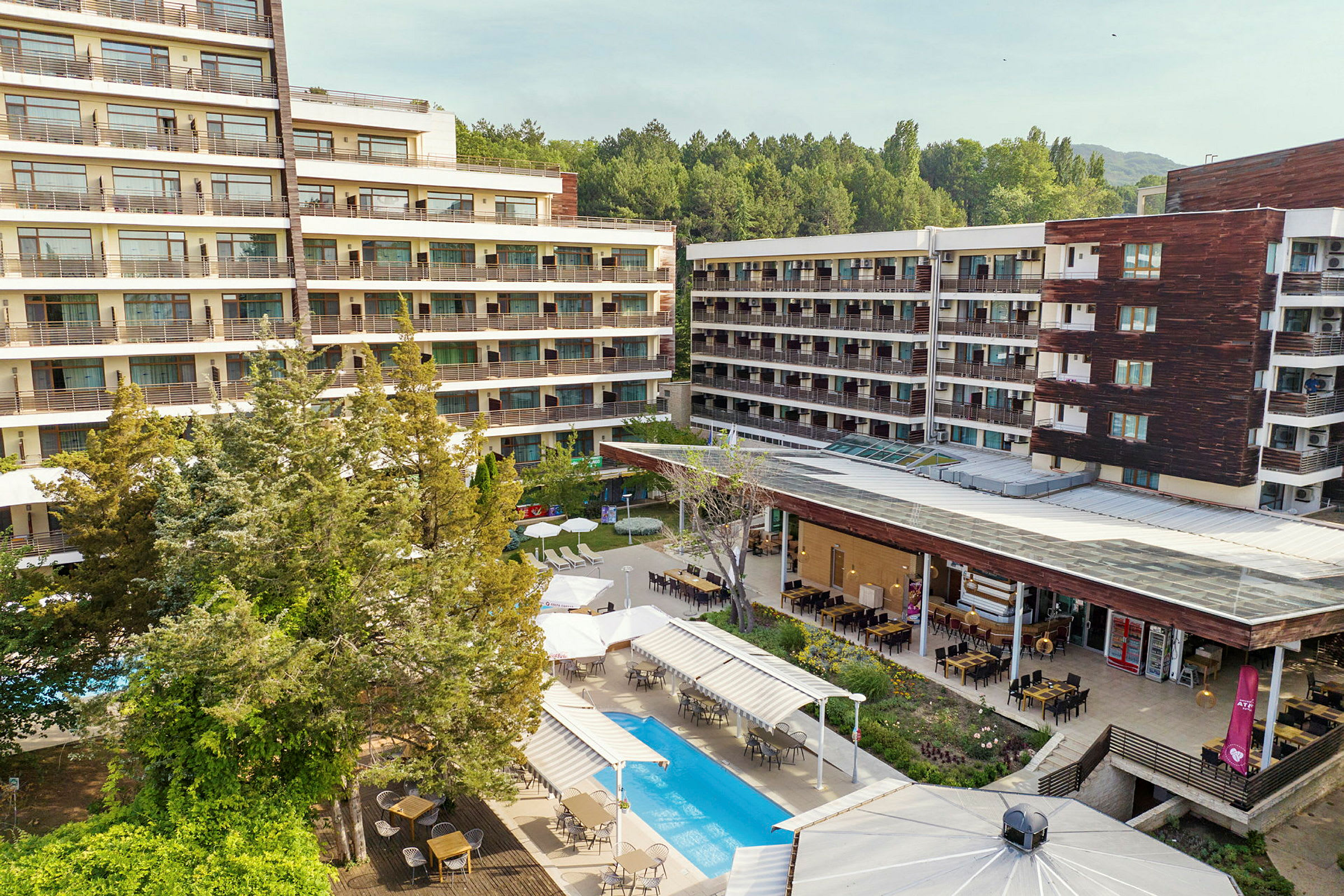 Hotellirakennuksia, joita ympäröi vehreys. Uima-allas ja ulkoruokailutila näkyvät etualalla. (Tekoälyn luoma teksti)
