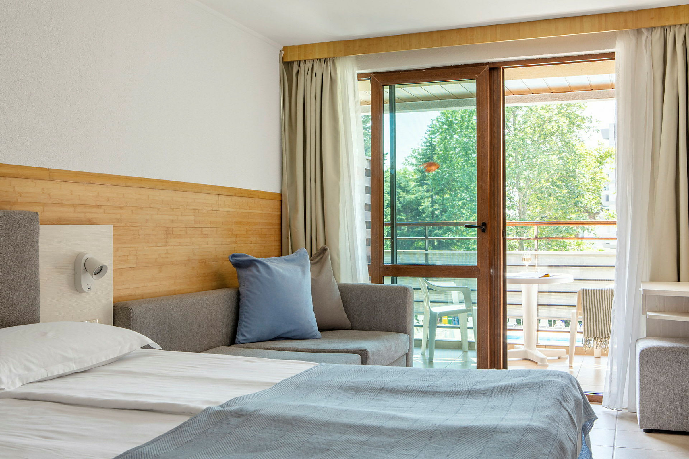 Hotellihuone, jossa on sänky, sohva ja parvekekalusteet näkyvissä lasioven läpi. (Tekoälyn luoma teksti)
