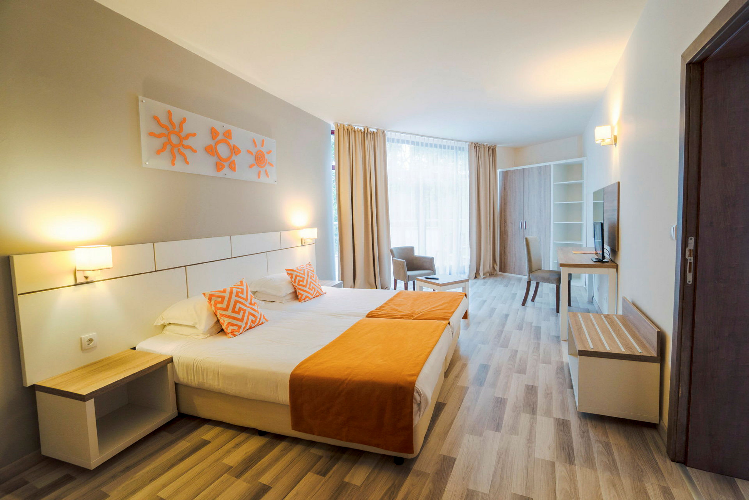 Hotellihuone, jossa on parisänky oransseilla yksityiskohdilla, kaksi sängyn valaisinta ja moderni sisustus. Leveä ikkuna ja vaaleat verhot näkyvät taustalla. (Tekoälyn luoma teksti)