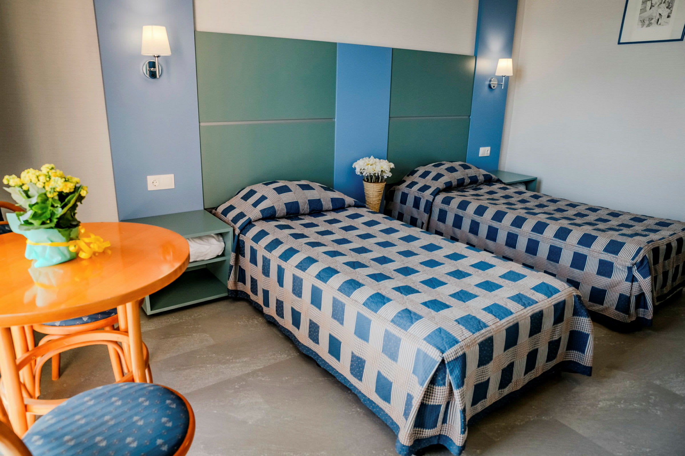 Hotellihuone, jossa kaksi yhden hengen sänkyä sinikuvioisilla päiväpeitteillä, pyöreä puinen pöytä tuolineen ja koristuksia yöpöydällä. (Tekoälyn luoma teksti)