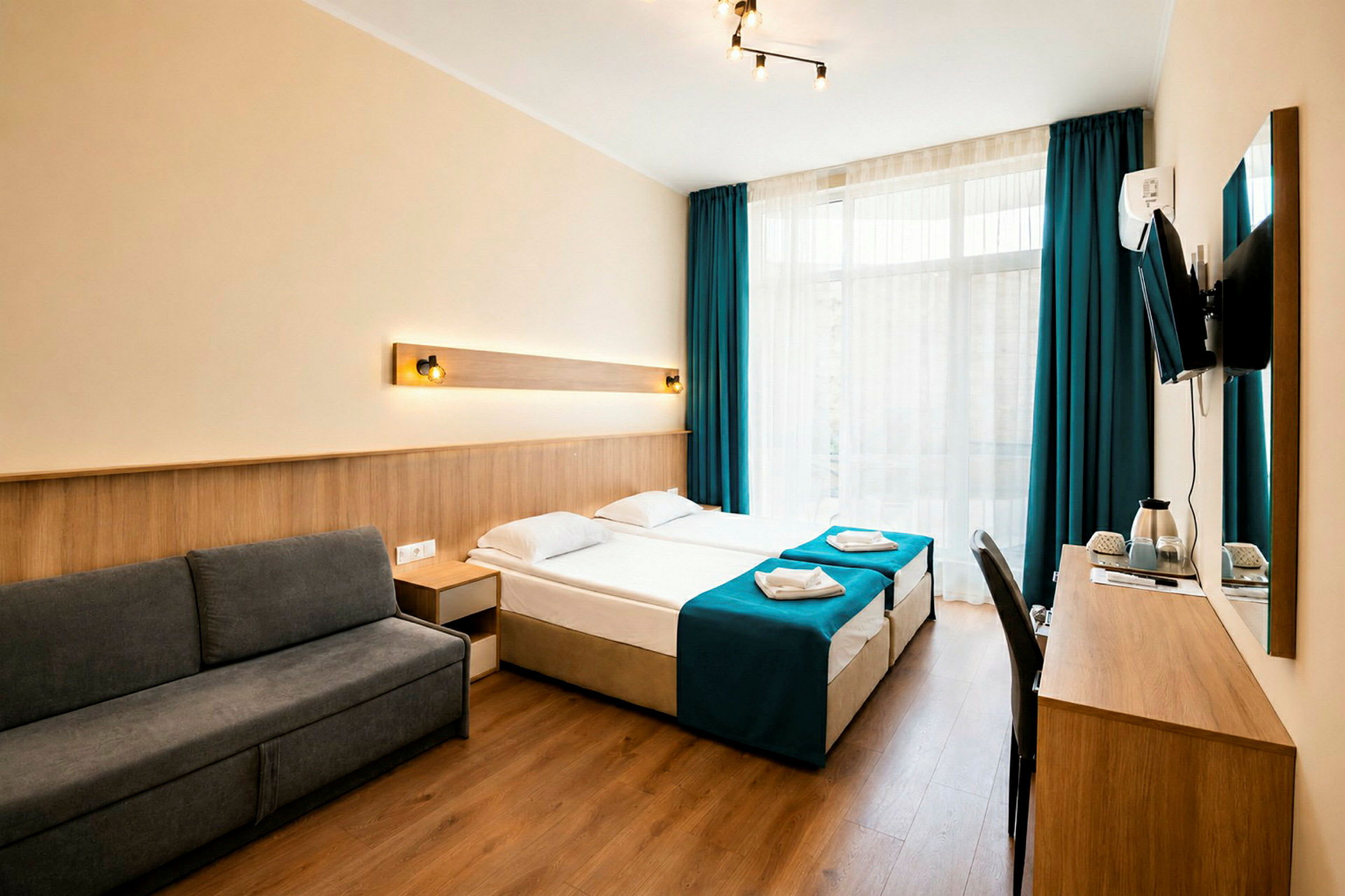 Hotellihuone, jossa on moderni sisustus, tummanharmaa sohva, kaksi erillistä vuodetta sinisillä päiväpeitteillä ja kirjoituspöytä seinään kiinnitetyllä televisiolla. (Tekoälyn luoma teksti)