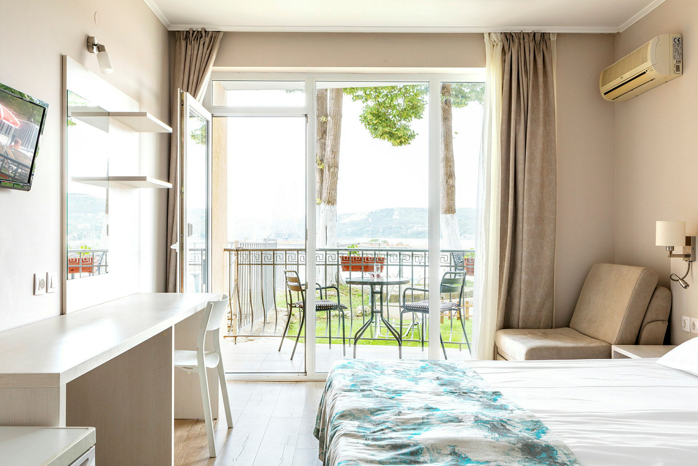 Hotellihuone, jossa on sänky, pöytä ja tuoli, sekä parveke ulkokalusteineen ja näkymä luontoon. (Tekoälyn luoma teksti)