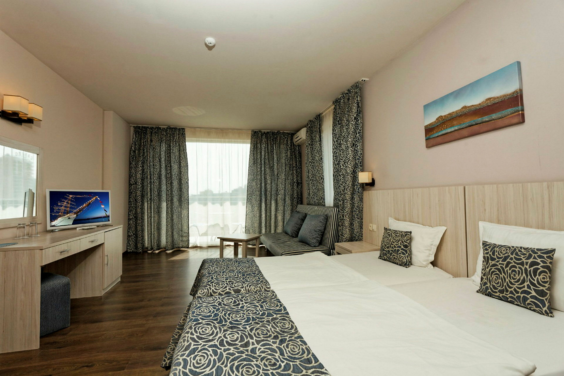 Hotellihuone, jossa on kaksi sänkyä, istuma-alue, televisio ja suuret ikkunat läpikuultavilla verhoilla. (Tekoälyn luoma teksti)