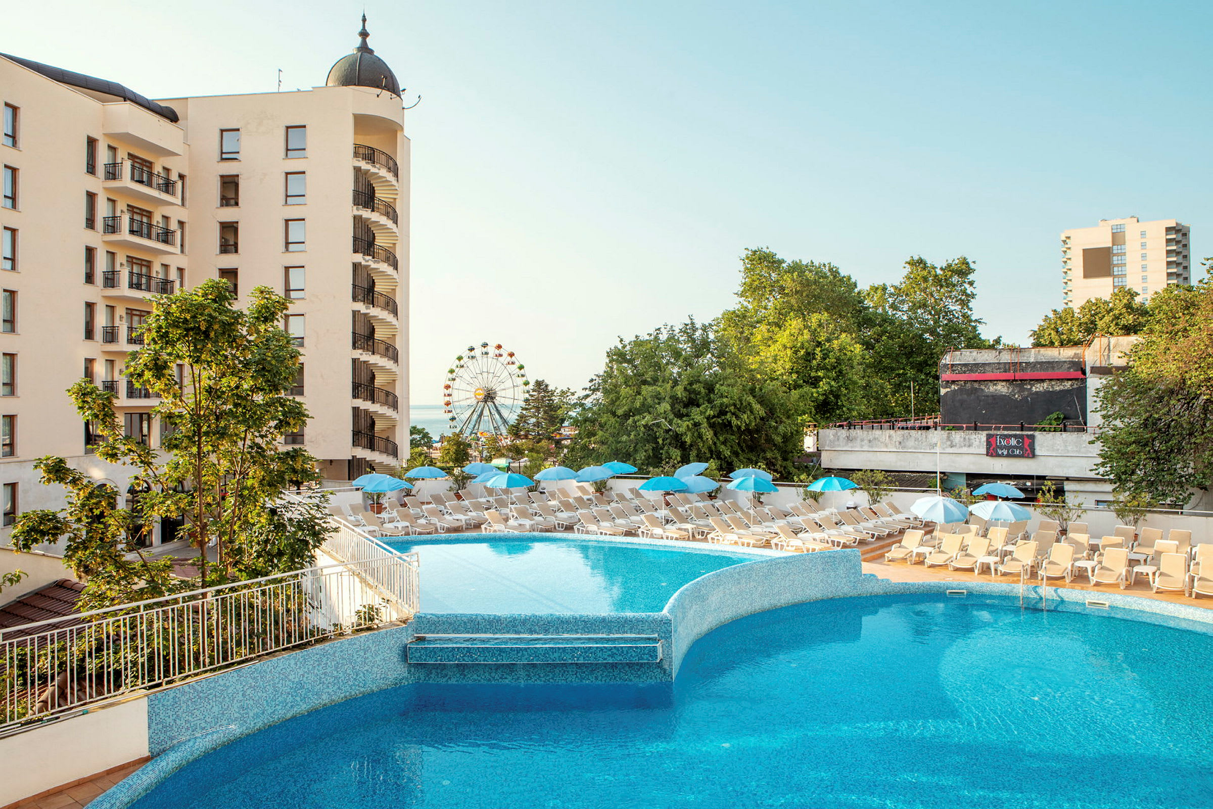 Hotellirakennus parvekkeineen, ulkouima-allas ja aurinkotuoleja varjoineen. Taustalla näkyy vihreitä puita ja maailmanpyörä. (Tekoälyn luoma teksti)