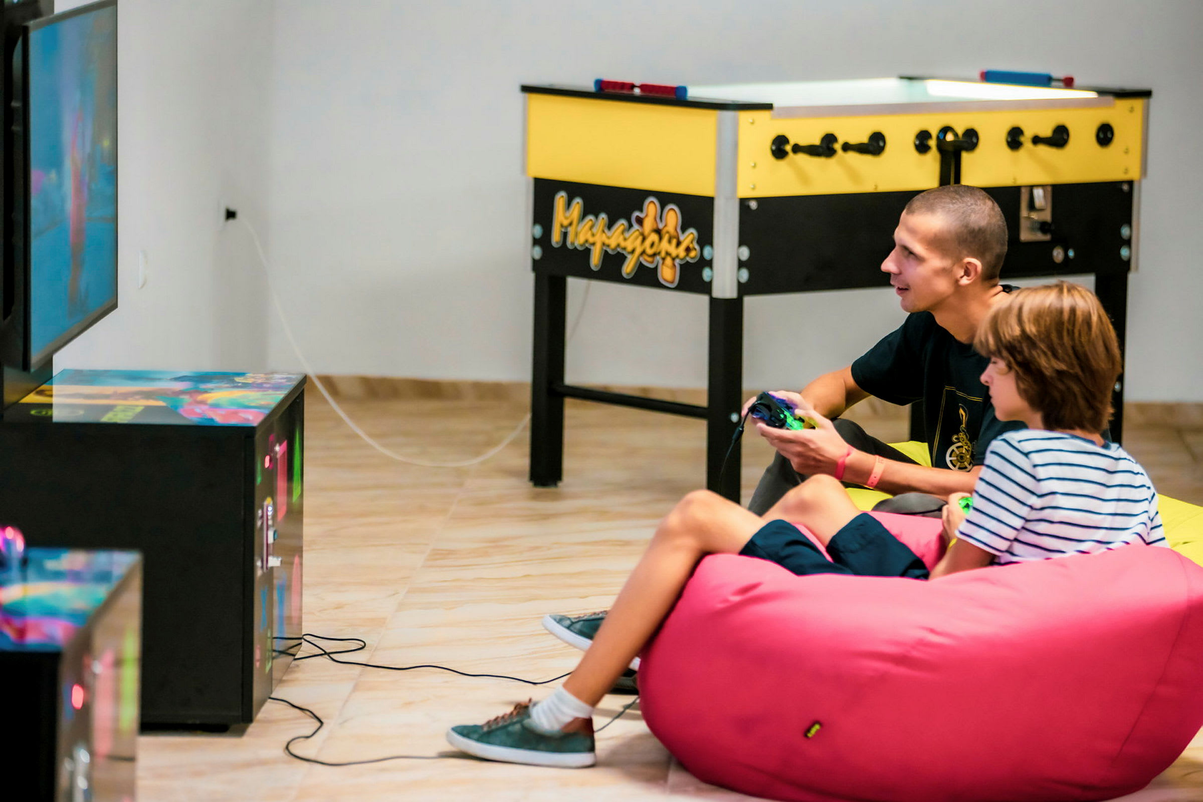 Kaksi henkilöä pelaa televisiopeliä istuen pinkillä säkkituolilla. Taustalla näkyy pöytäjalkapallopeli. (Tekoälyn luoma teksti)