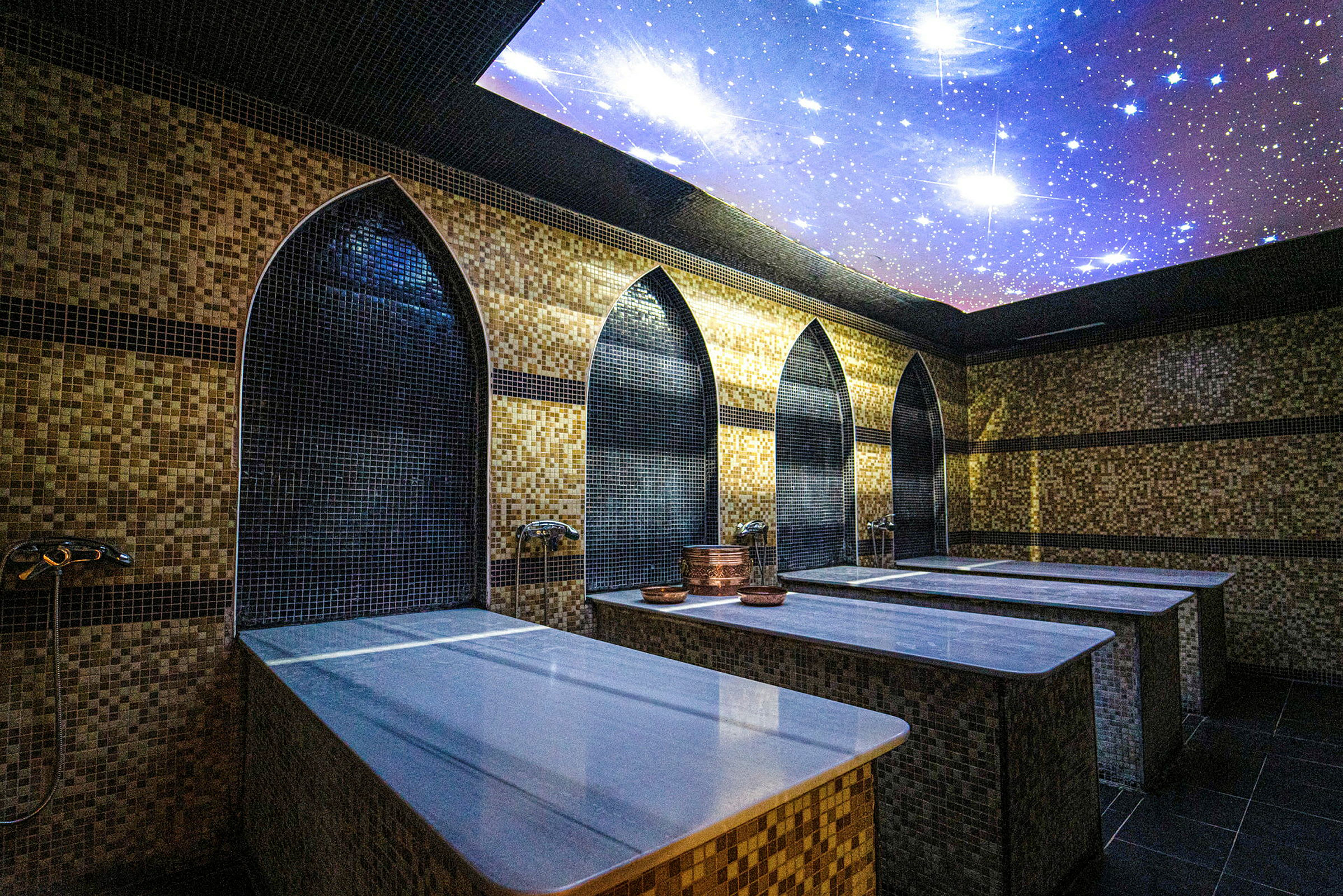 Huoneessa näkyy perinteinen hamam marmoripöytineen ja koristeellisilla mosaiikkiseinillä. Katossa on tähdillä koristeltu kuvio. (Tekoälyn luoma teksti)