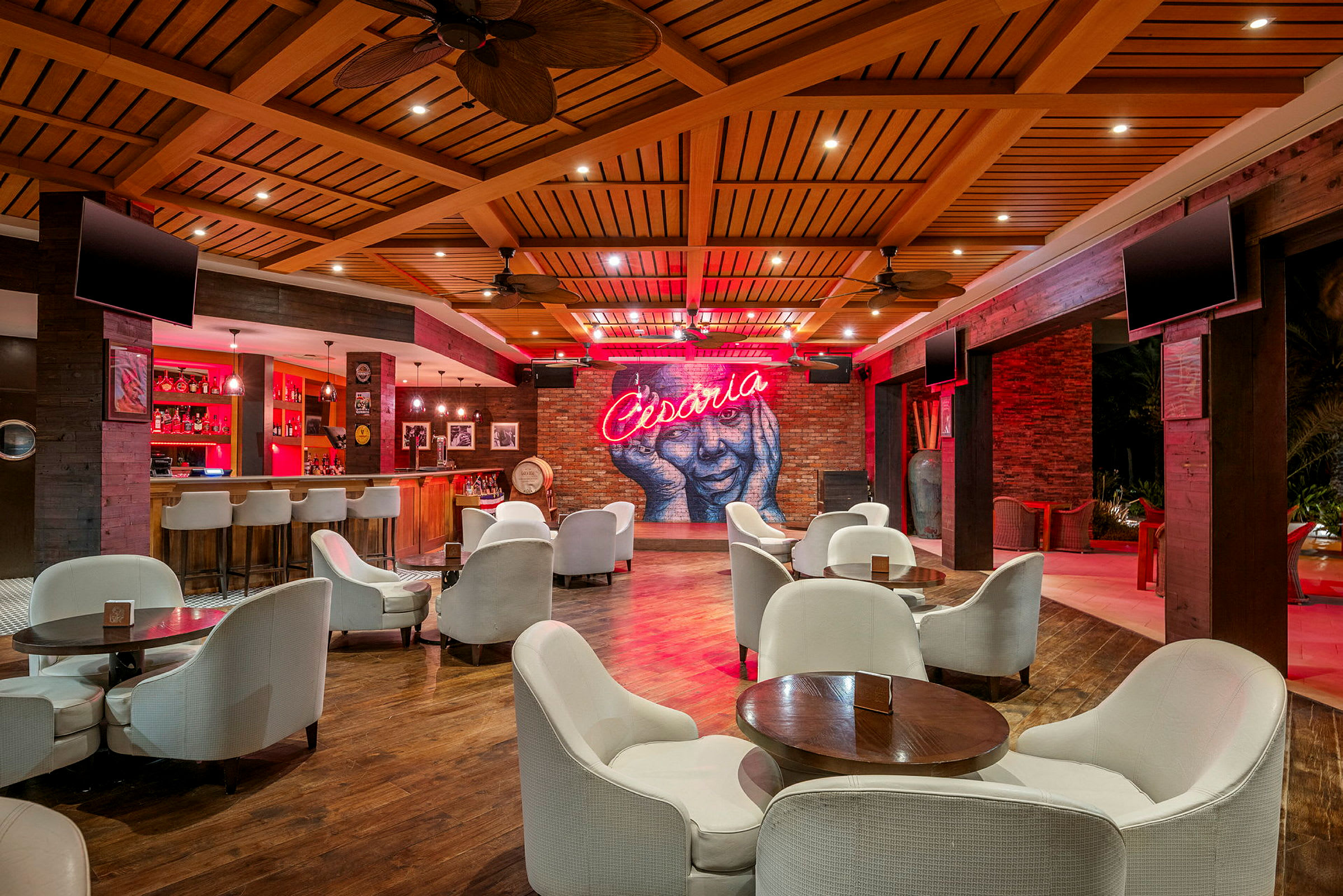 Tyylikäs lounge, jossa on valkoisia tuoleja ja pöytiä, taustalla baari ja seinällä neonteksti 'Cesaria'. (Tekoälyn luoma teksti)