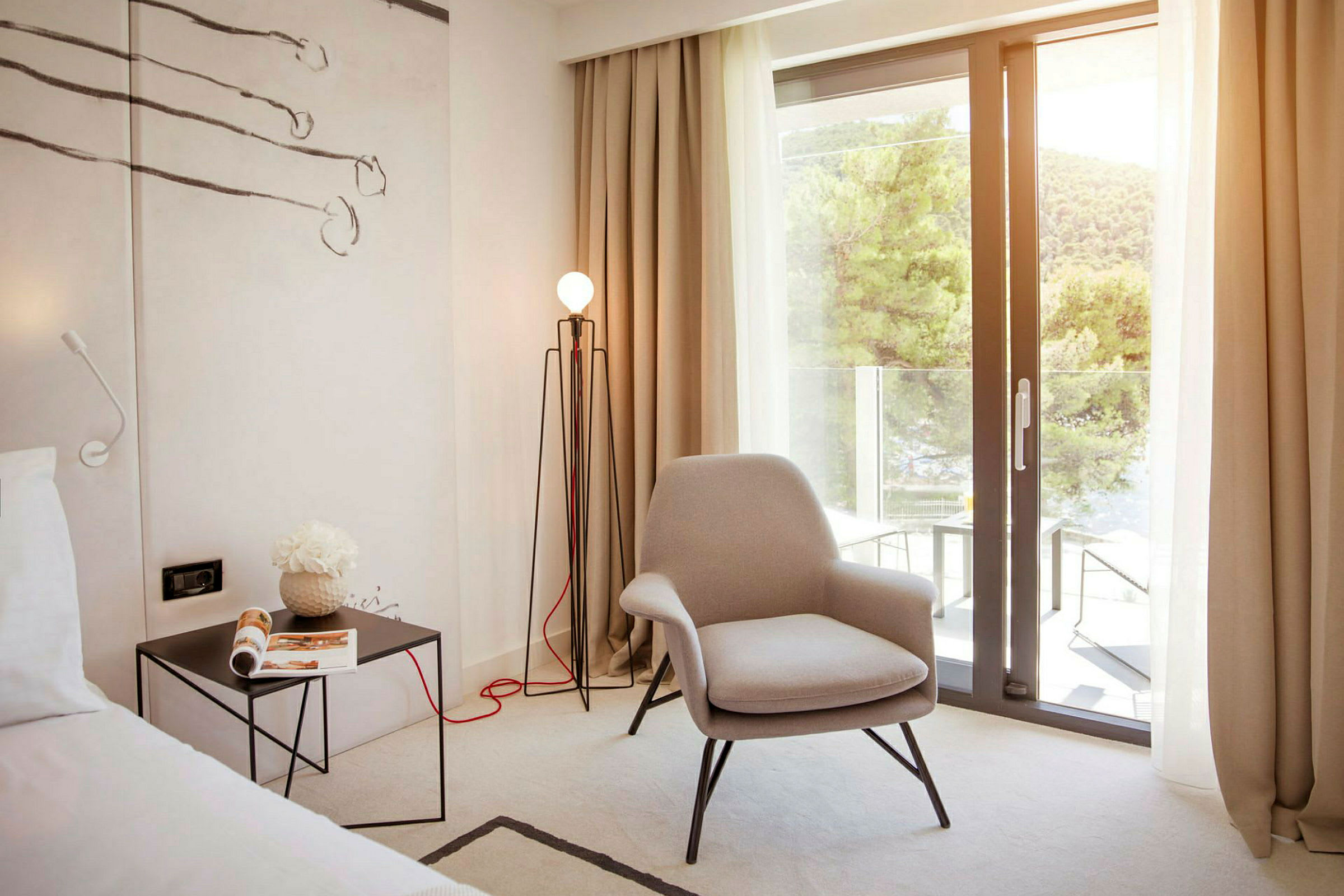 Hotellihuone, jossa on mukava nojatuoli, sivupöytä, jossa on lehti, ja lattiavalaisin. Suuri lasiovi avautuu parvekkeelle, josta on näköala vehreisiin puihin. (Tekoälyn luoma teksti)