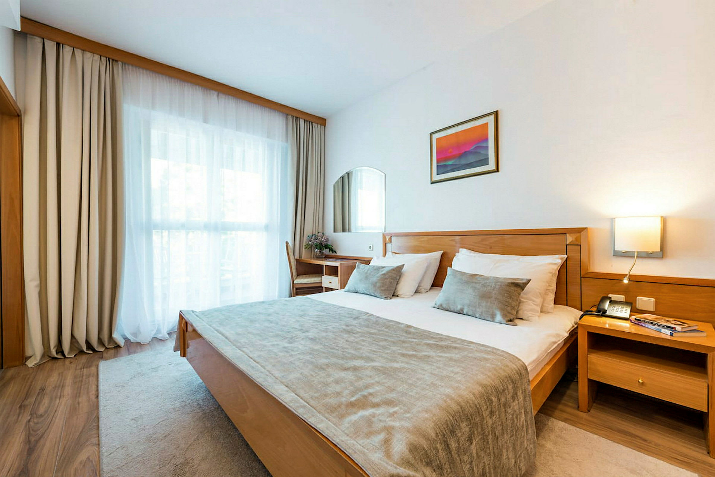 Hotellihuone, jossa on parivuode, kirjoituspöytä tuoleineen, ikkuna verhoilla ja seinäkoriste. (Tekoälyn luoma teksti)