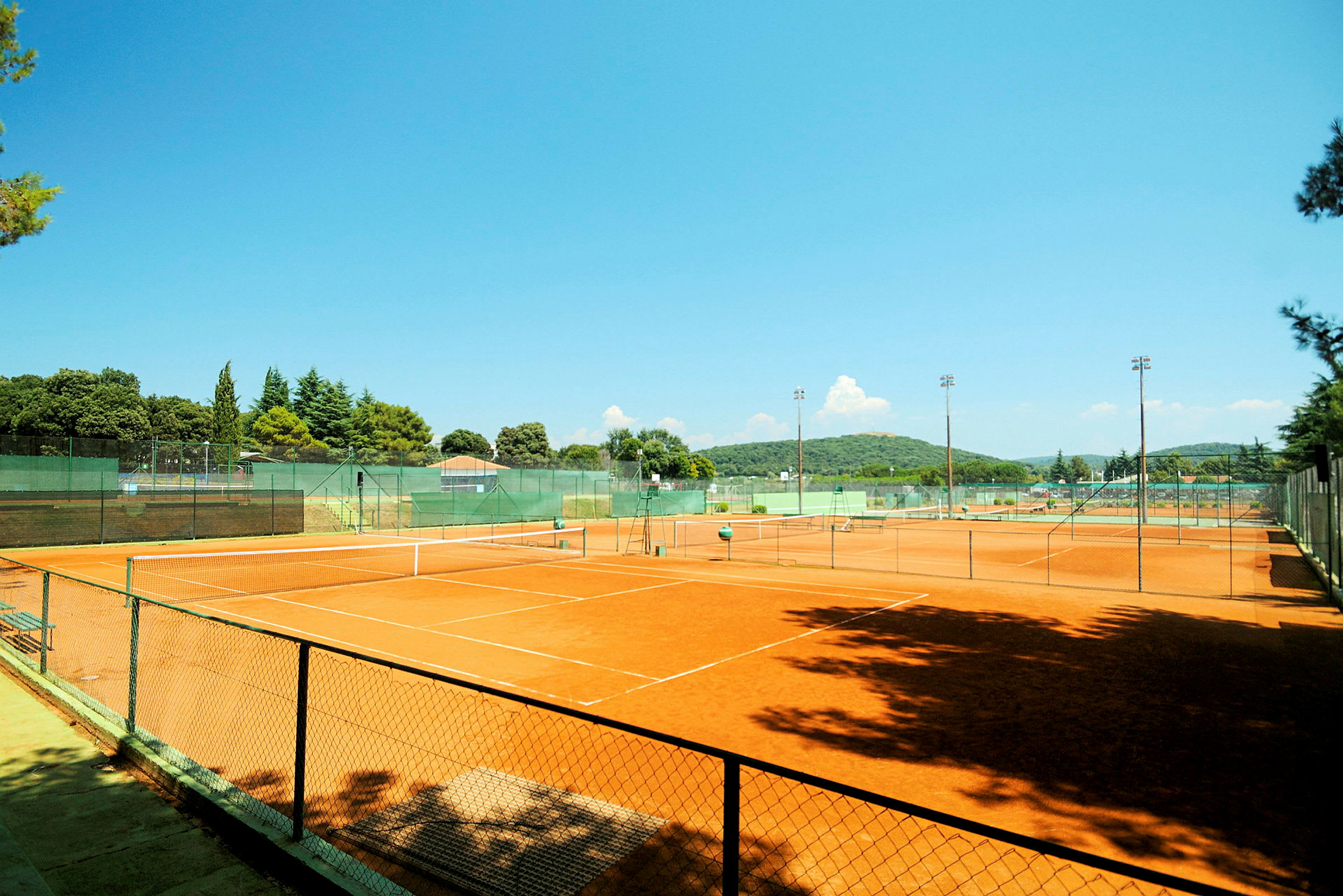 Kuva ulkotenniskentästä sora-alustalla, jota ympäröi vihreys ja aita. (Tekoälyn luoma teksti)