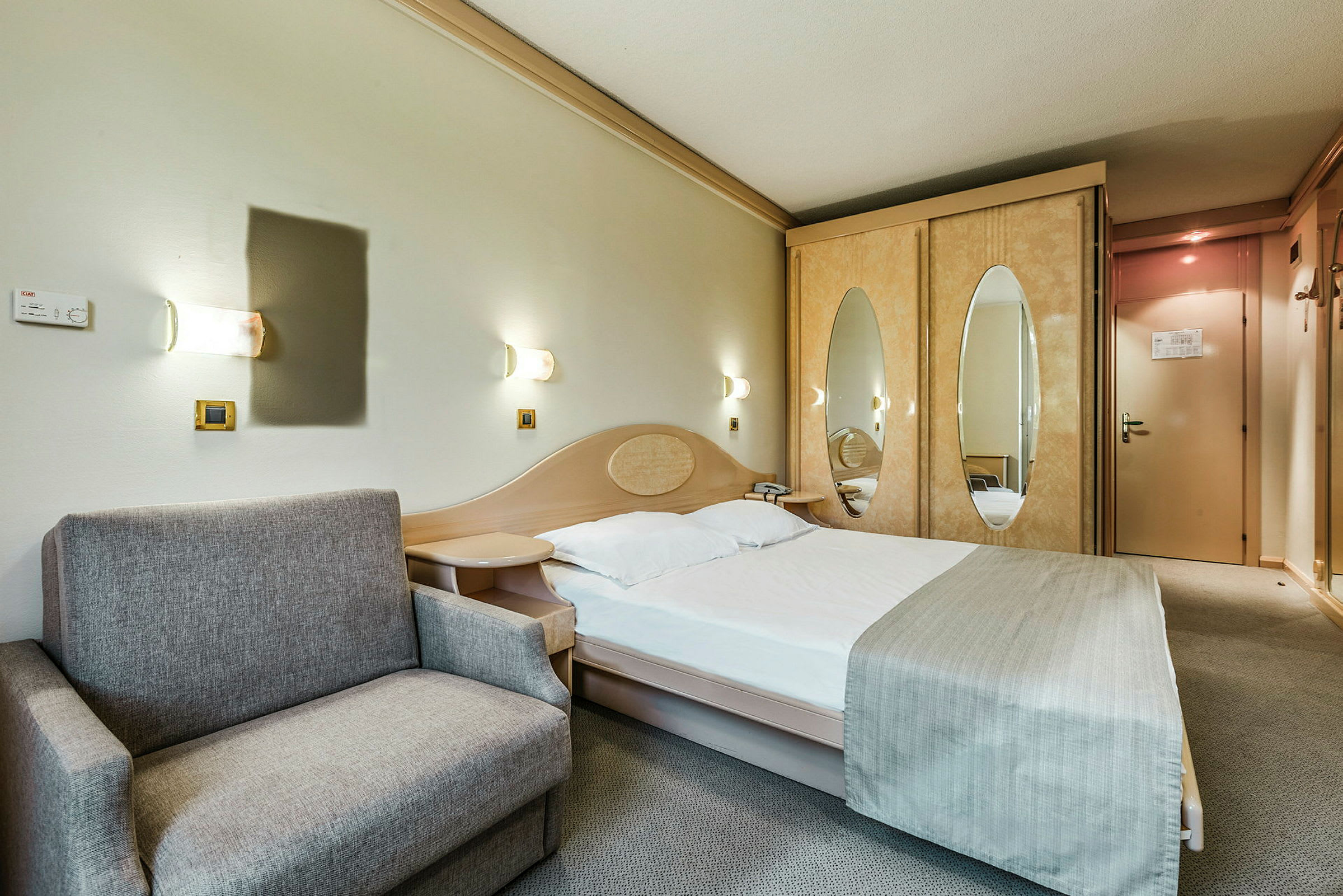 Hotellihuone, jossa on pedattu sänky, harmaa sohva ja vaatekaappi, jossa on peiliovet. (Tekoälyn luoma teksti)