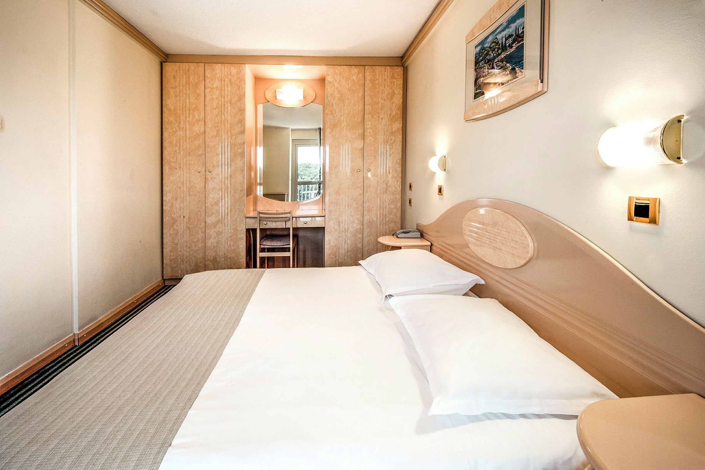 Hotellihuone, jossa on parisänky, seinävalaisimia ja sisäänrakennettu vaatekaappi, jonka edessä on peili ja tuoli. (Tekoälyn luoma teksti)