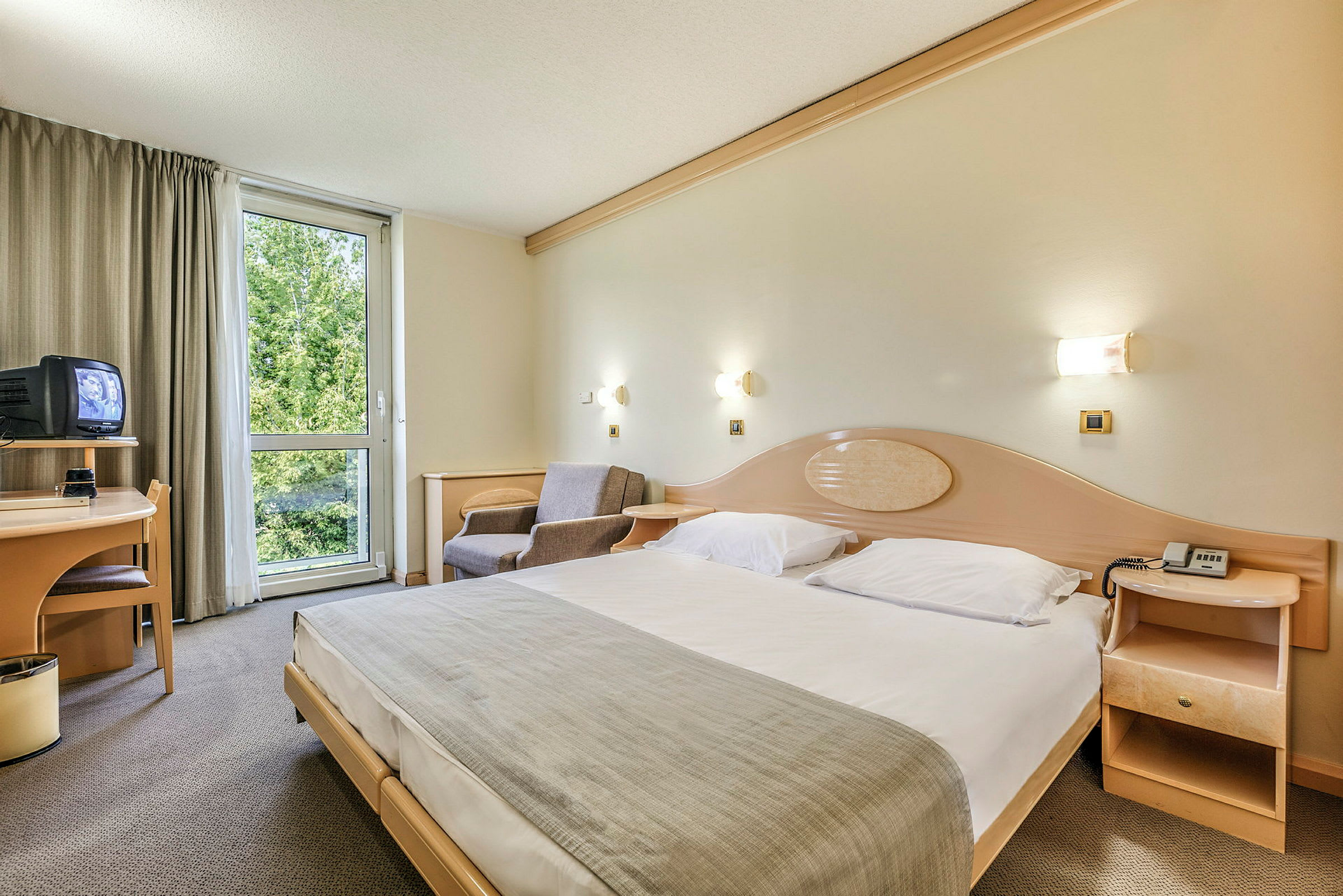 Hotellihuone, jossa on suuri sänky, nojatuoli, kirjoituspöytä, televisio ja ikkuna, josta on näkymä vehreyteen. (Tekoälyn luoma teksti)