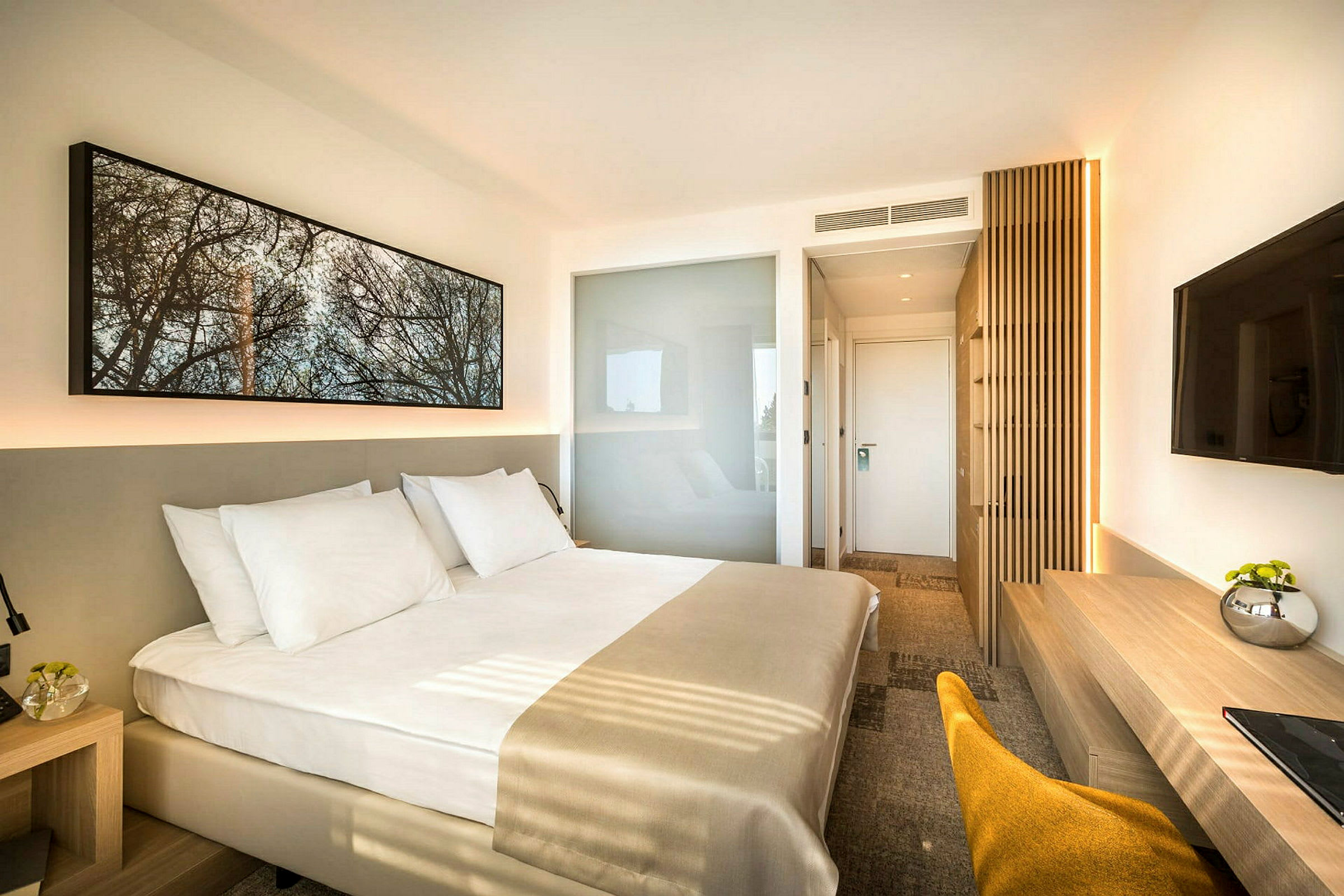 Moderni hotellihuone, jossa on suuri sänky valkoisilla vuodevaatteilla. Huoneessa on minimalistinen sisustus, seinälle kiinnitetty televisio, työpöytä ja tuoli. (Tekoälyn luoma teksti)