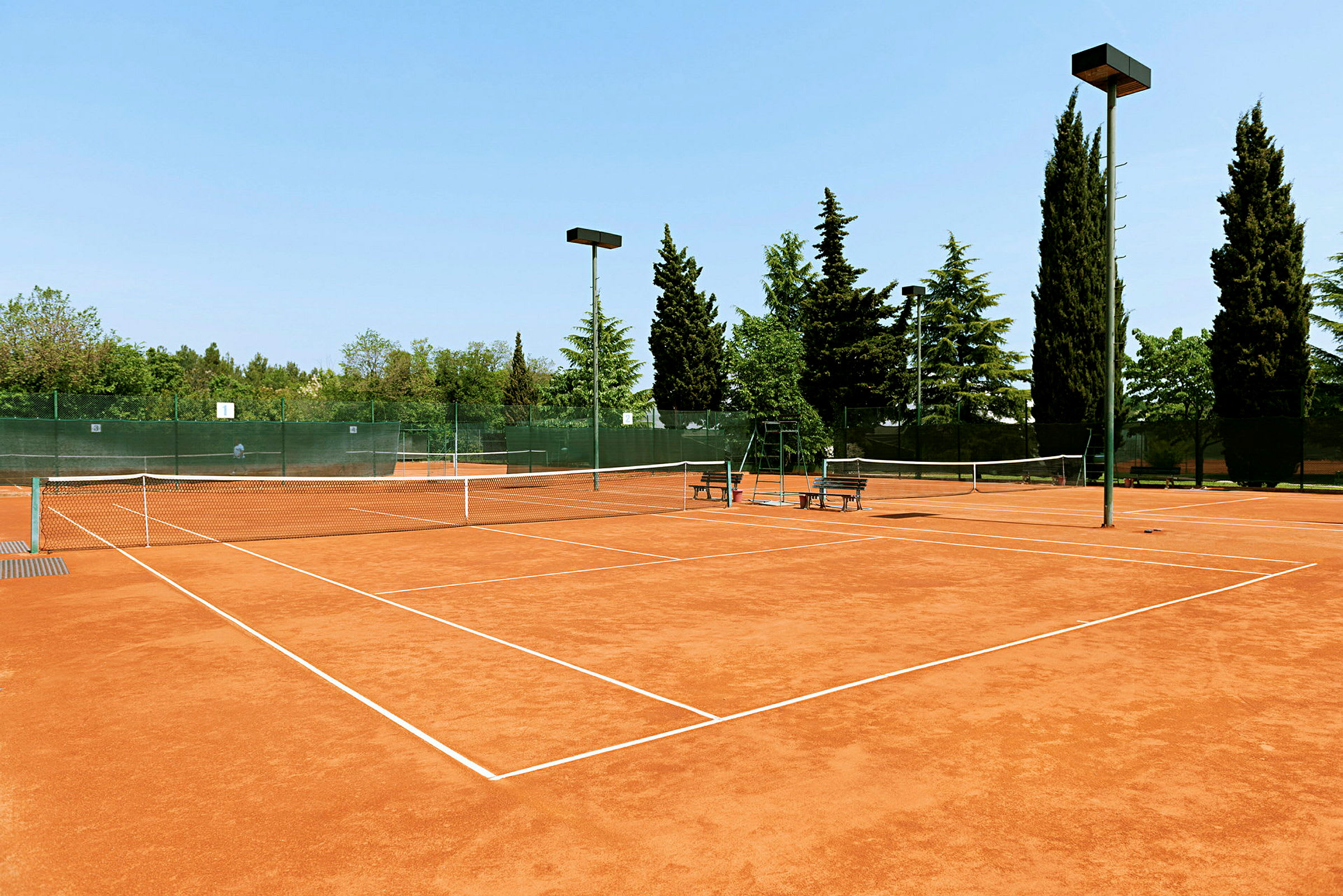 Savikenttäinen tenniskenttä, jota ympäröivät puut ja aita. (Tekoälyn luoma teksti)