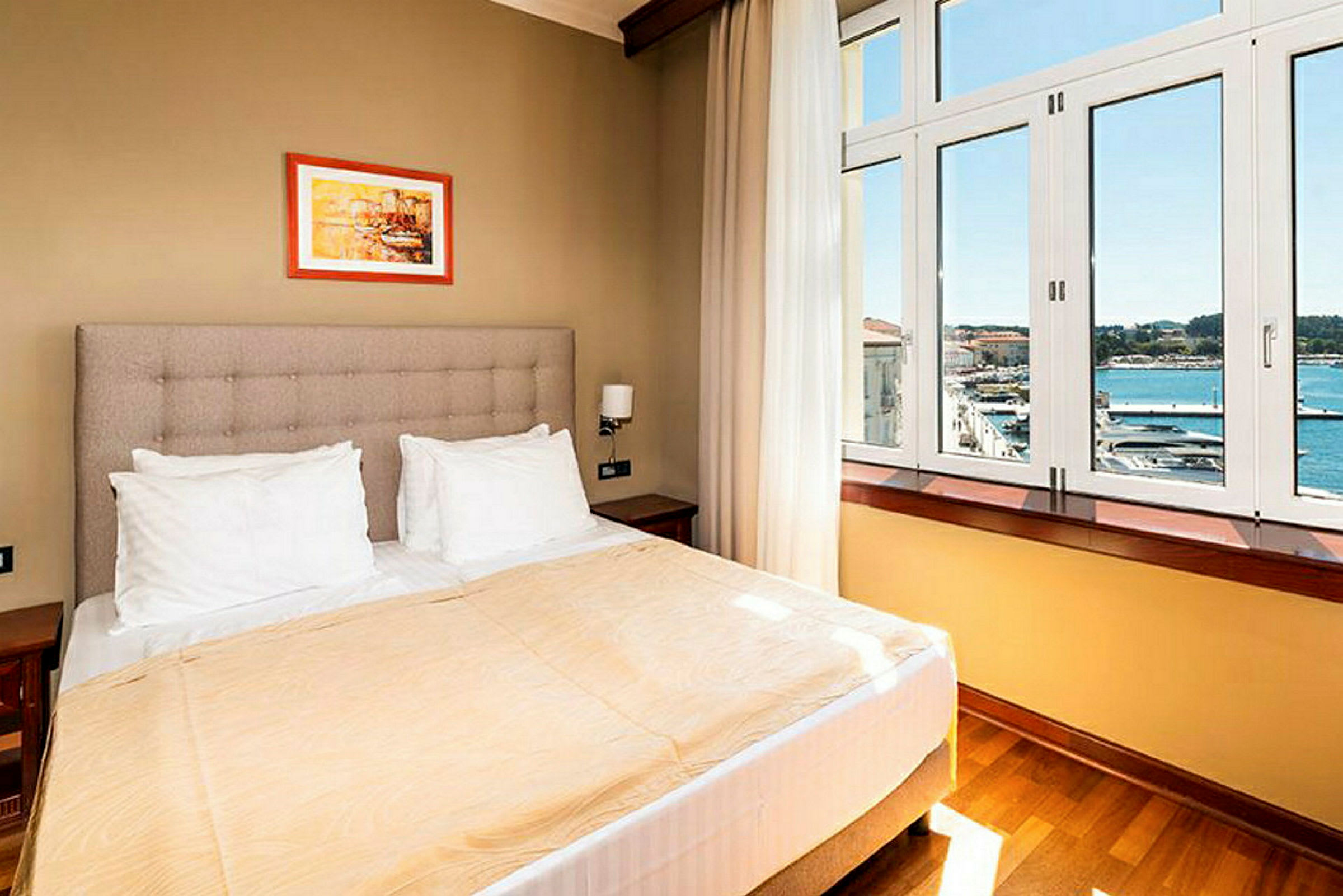 Hotellihuone, jossa on parivuode, valkoiset tyynyt, beige päiväpeite ja iso ikkuna, josta näkyy merimaisema. (Tekoälyn luoma teksti)