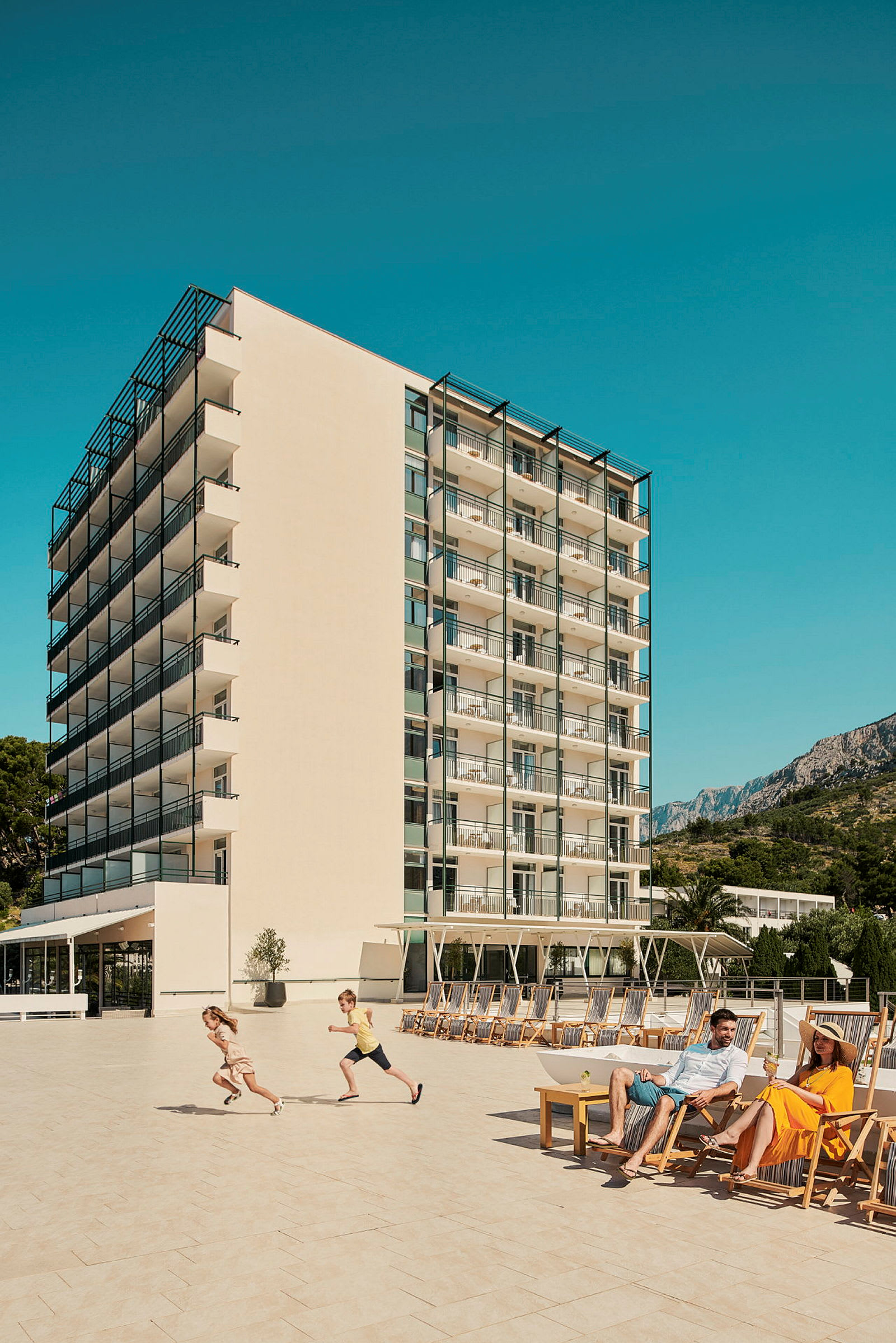 Kuva hotellista, jossa on parvekkeita useissa kerroksissa, suuri terassi aurinkotuoleineen ja ihmisiä nauttimassa auringosta. (Tekoälyn luoma teksti)