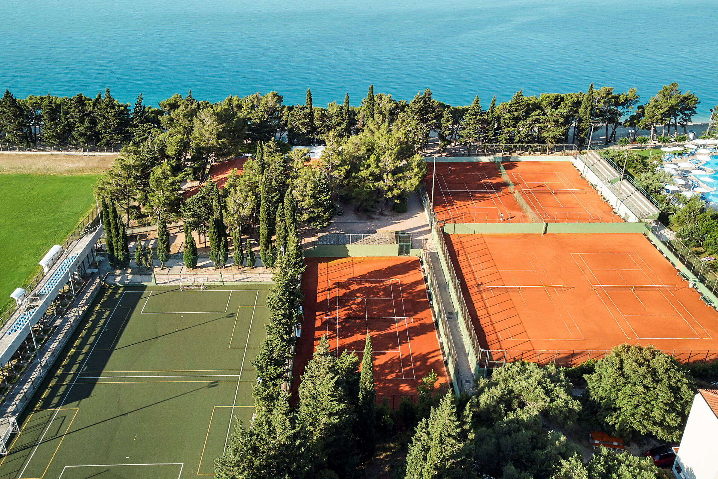 Ilmakuva urheilutilasta, jossa on tenniskenttiä ja jalkapallokenttä, ympäröity puilla ja merellä. (Tekoälyn luoma teksti)