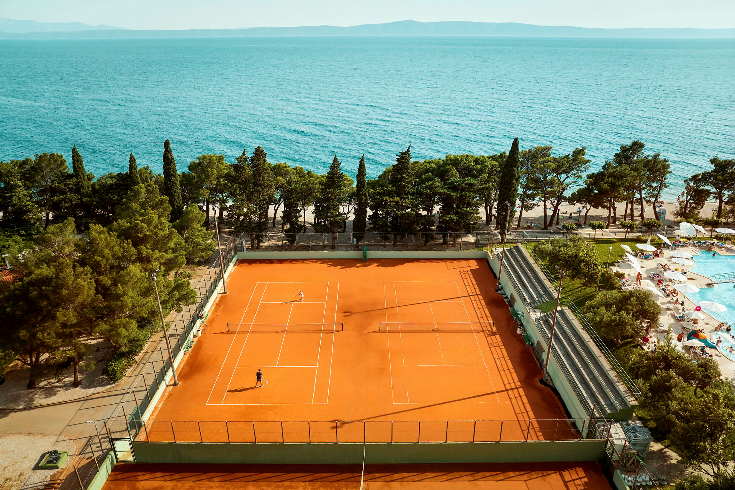 Tenniskenttä pelaajineen lähellä merta ja uima-allasaluetta. (Tekoälyn luoma teksti)
