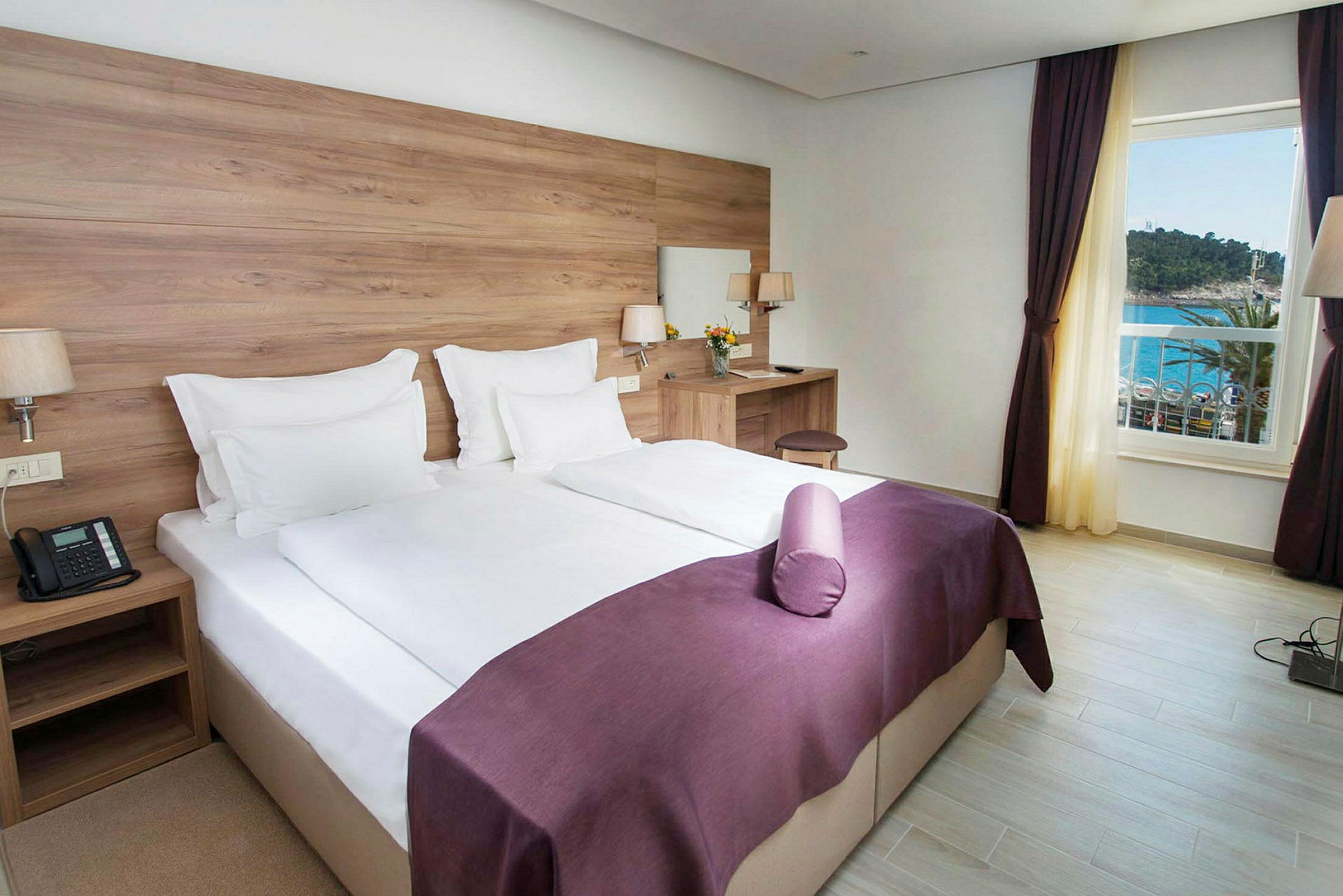 Hotellihuone, jossa on parivuode, valkoiset lakanat ja violetti päiväpeitto. Huoneessa on kirjoituspöytä tuolilla ja peili. Ikkunasta on näkymä merelle. (Tekoälyn luoma teksti)