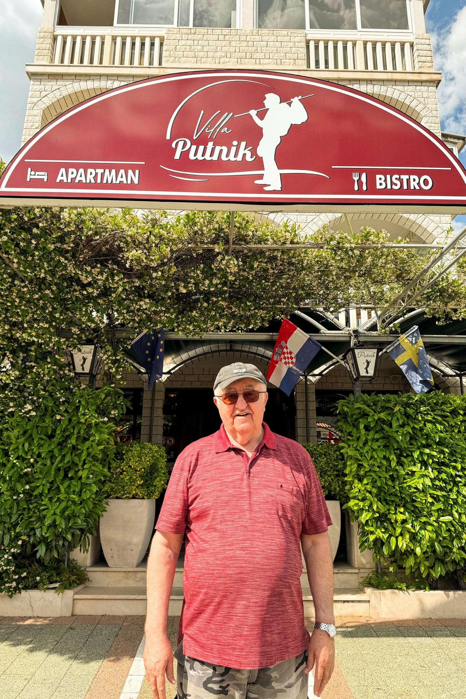 Kuva henkilöstä, joka seisoo rakennuksen edessä. Rakennuksessa on kyltti, jossa lukee "Villa Putnik", ja ympärillä on vihreitä kasveja. (Tekoälyn luoma teksti)