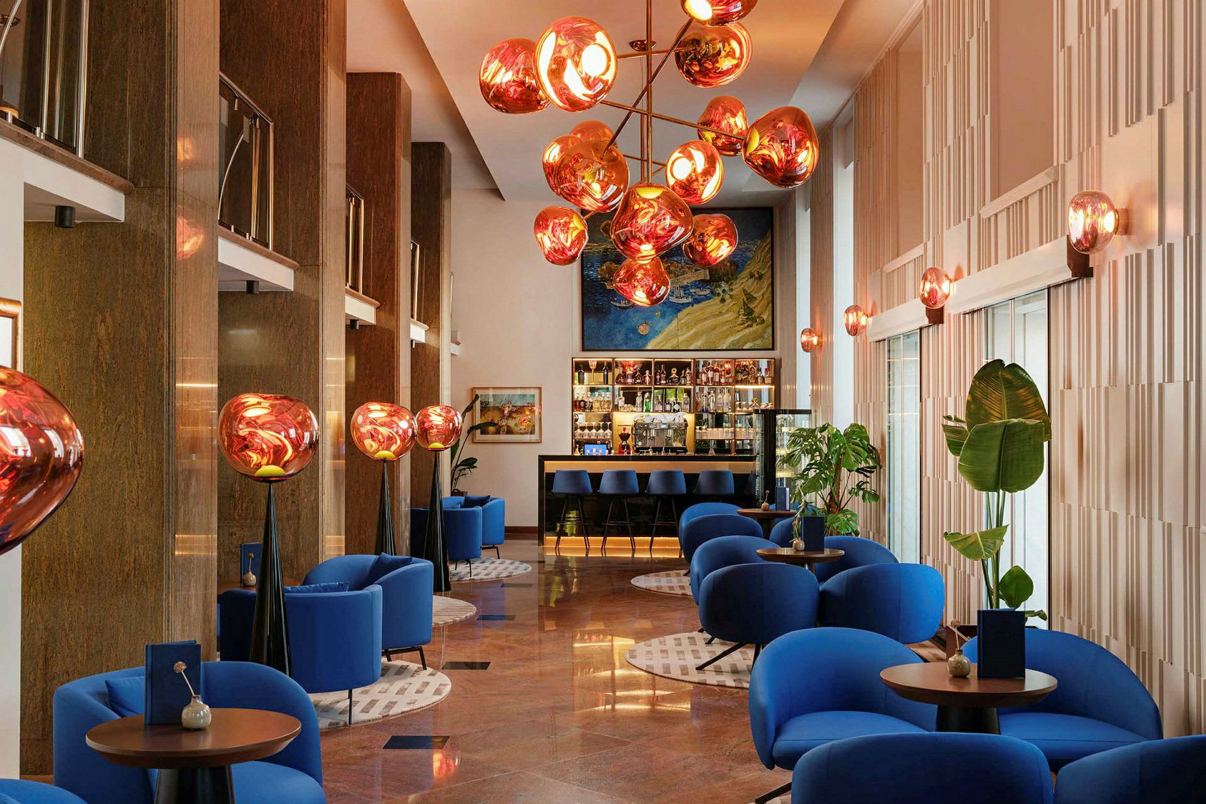 Moderni hotelliaula, jossa siniset nojatuolit, pyöreitä pöytiä ja koristeellinen kattovalaistus. (Tekoälyn luoma teksti)
