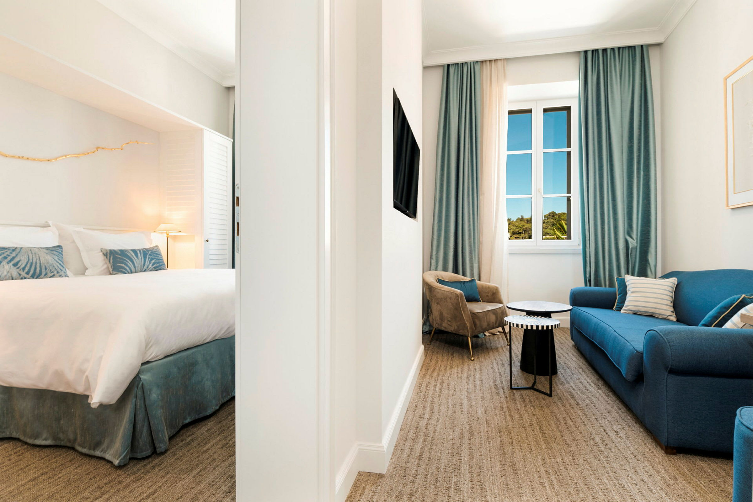 Hotellihuoneisto, jossa on makuuhuone ja olohuone, sisustettuna sinisellä sohvalla, nojatuoleilla ja siniset verhot omaavalla ikkunalla. (Tekoälyn luoma teksti)