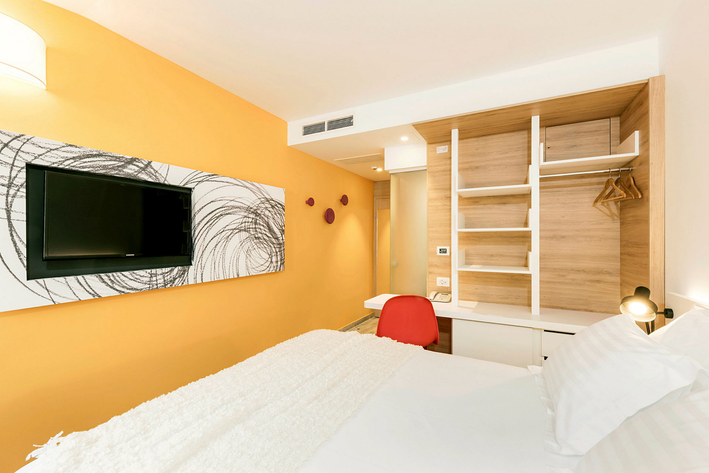 Hotellihuone, jossa on keltainen seinä, seinällä televisio, punainen tuoli pöydän ääressä ja sänky, jossa on valkoiset vuodevaatteet. (Tekoälyn luoma teksti)