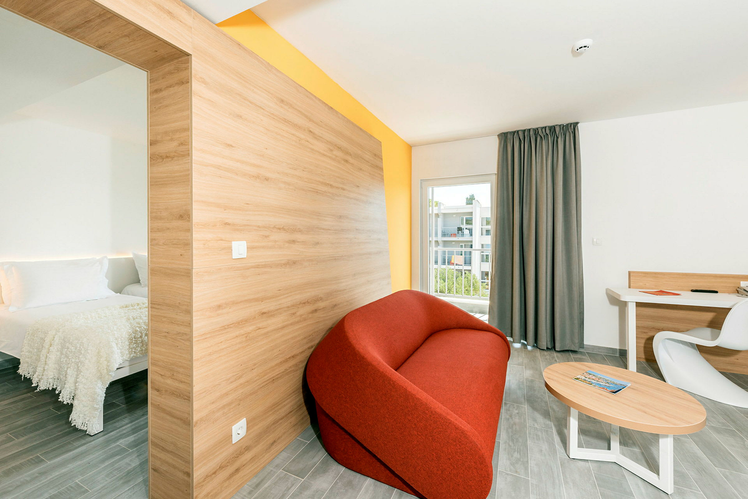 Moderni hotellihuone, jossa erillinen makuutila vasemmalla, punainen sohva, pöytä ja työtila tuolillaan sekä parveke näkymällä. (Tekoälyn luoma teksti)