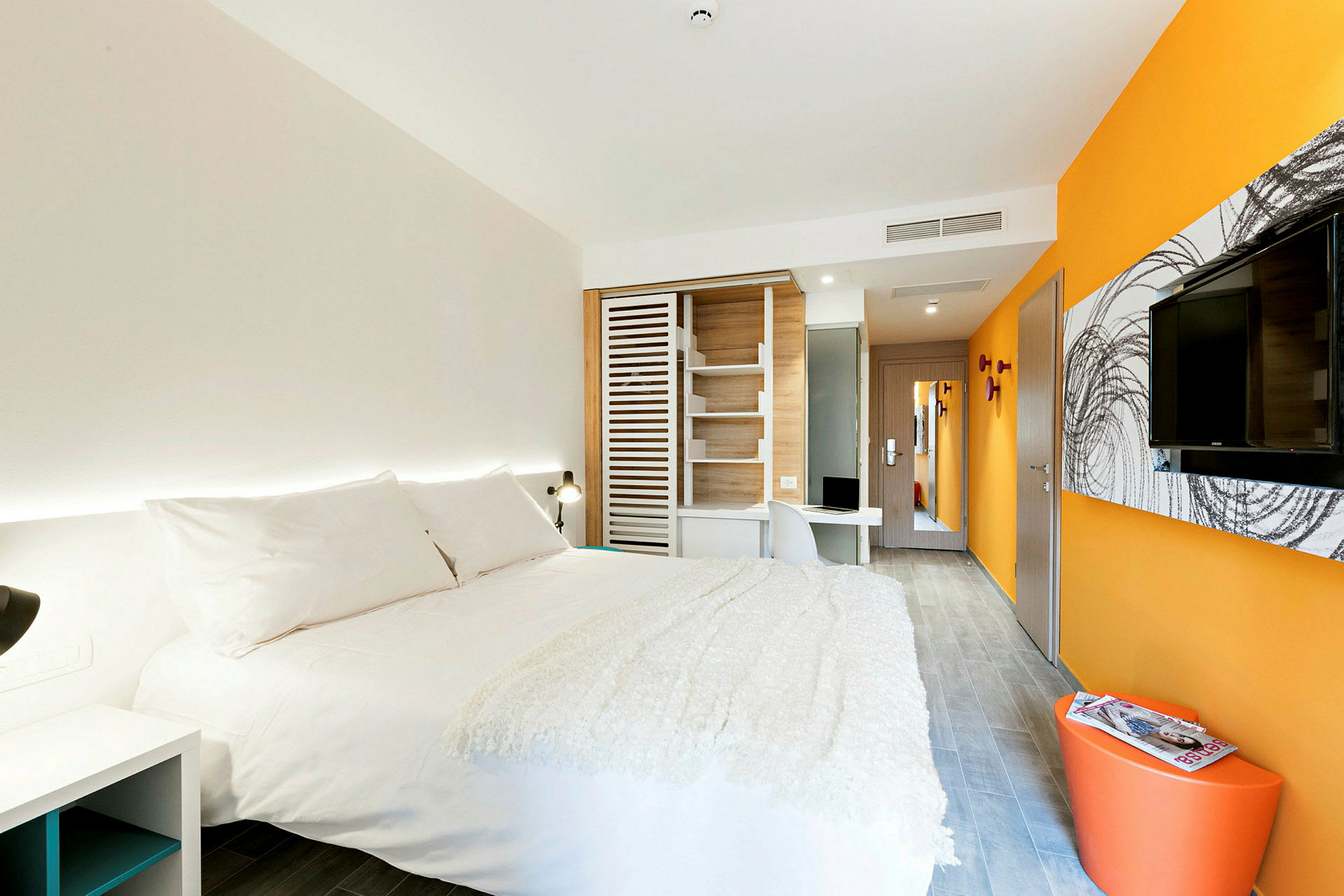 Moderni hotellihuone, jossa on suuri sänky valkoisilla vuodevaatteilla, oranssit ja valkoiset seinät, seinälle asennettu televisio ja työpöytä. (Tekoälyn luoma teksti)