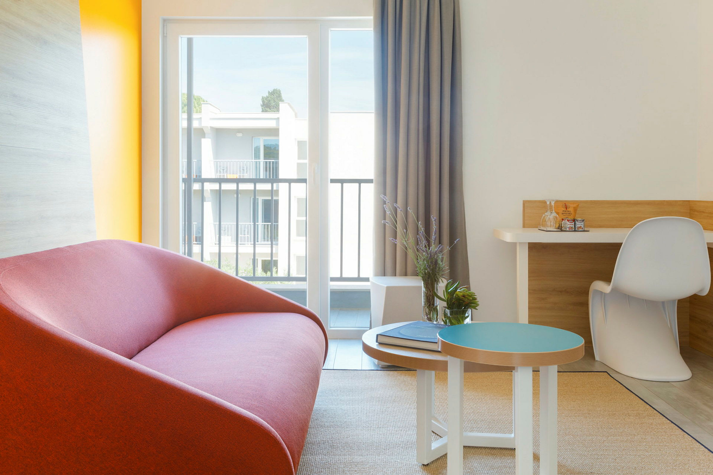 Modernisti sisustettu hotellihuoneen kulma, jossa on punainen sohva, pieni koristeellinen pöytä ja kirjoituspöytänurkkaus suuren ikkunan ääressä. (Tekoälyn luoma teksti)