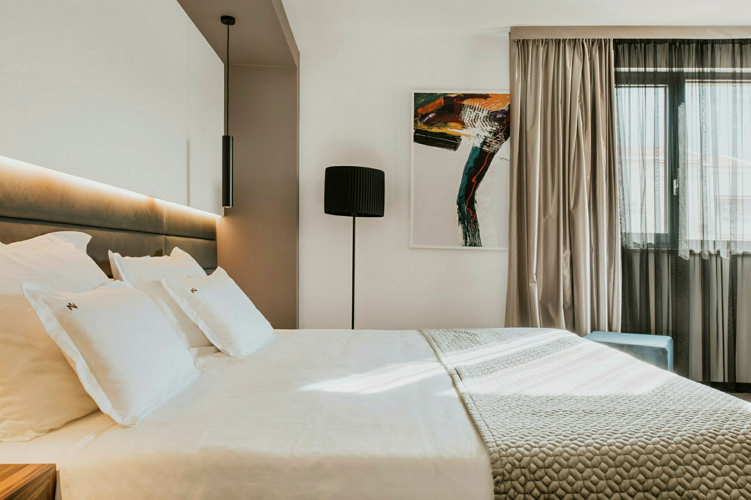 Moderni hotellihuone, jossa on suuri sänky, taideteos seinällä ja ikkuna kevyillä verhoilla. (Tekoälyn luoma teksti)