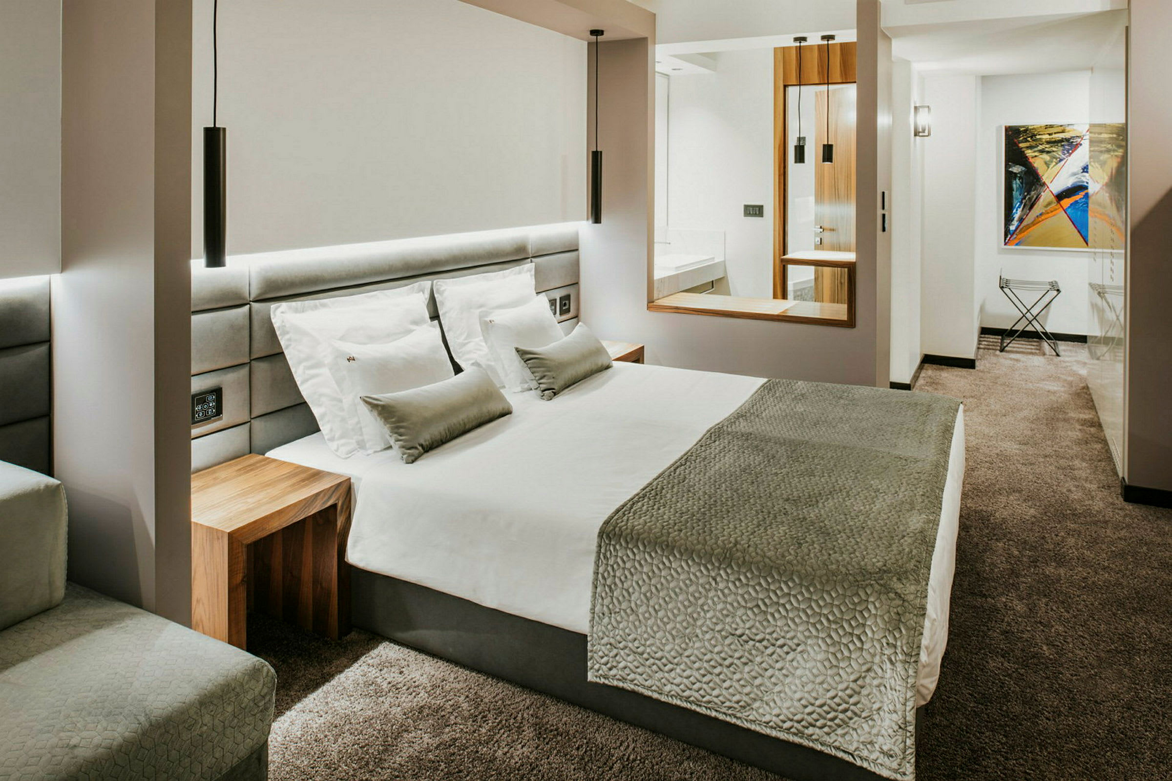 Moderni hotellihuone, jossa on suuri sänky, valkoinen vuodevaatteet, harmaa päiväpeite ja tyynyt sekä harmaa kokolattiamatto. (Tekoälyn luoma teksti)