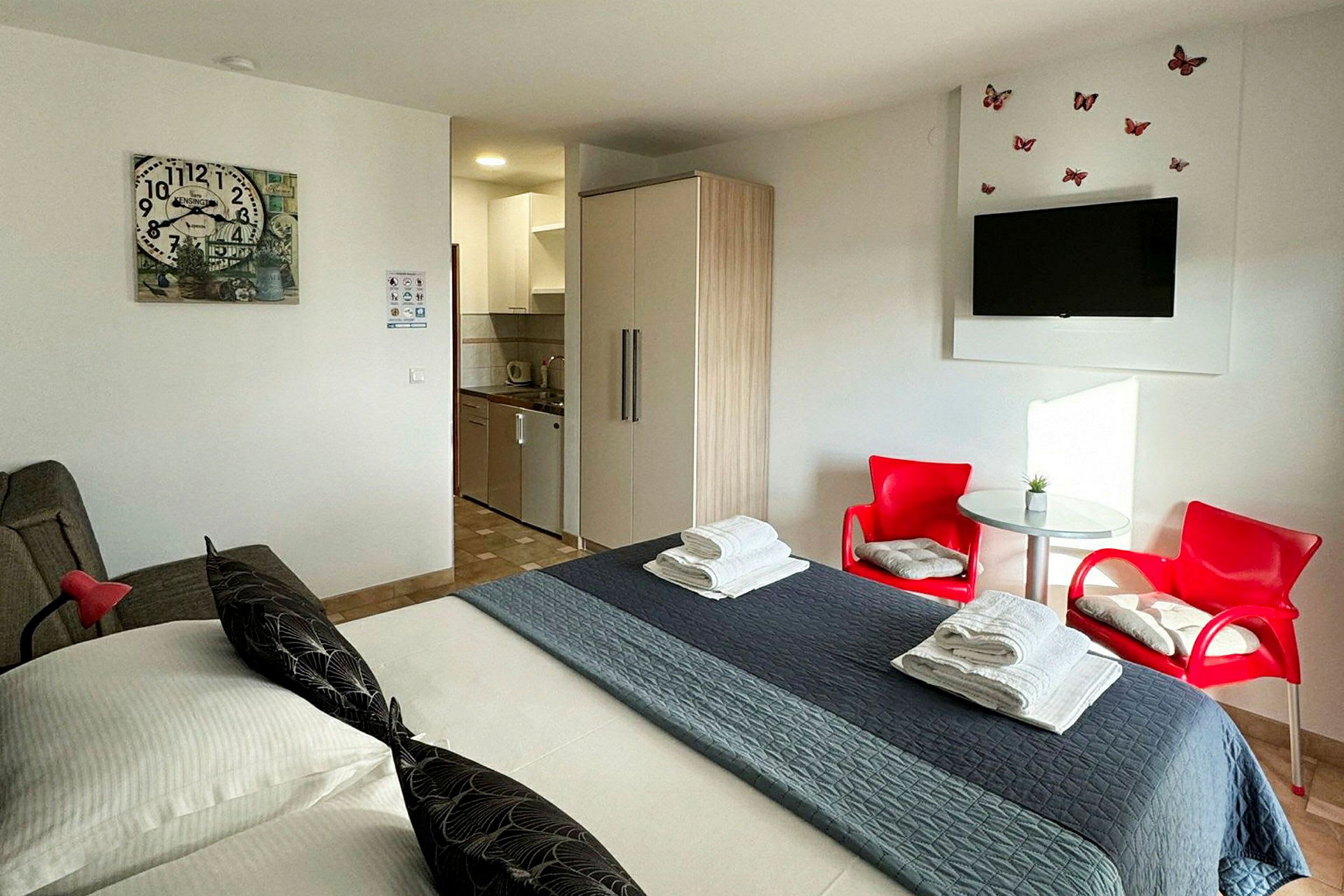 Hotellihuone, jossa on sänky, punaisia tuoleja, pöytä, vaatekaappi ja pieni keittiö. (Tekoälyn luoma teksti)