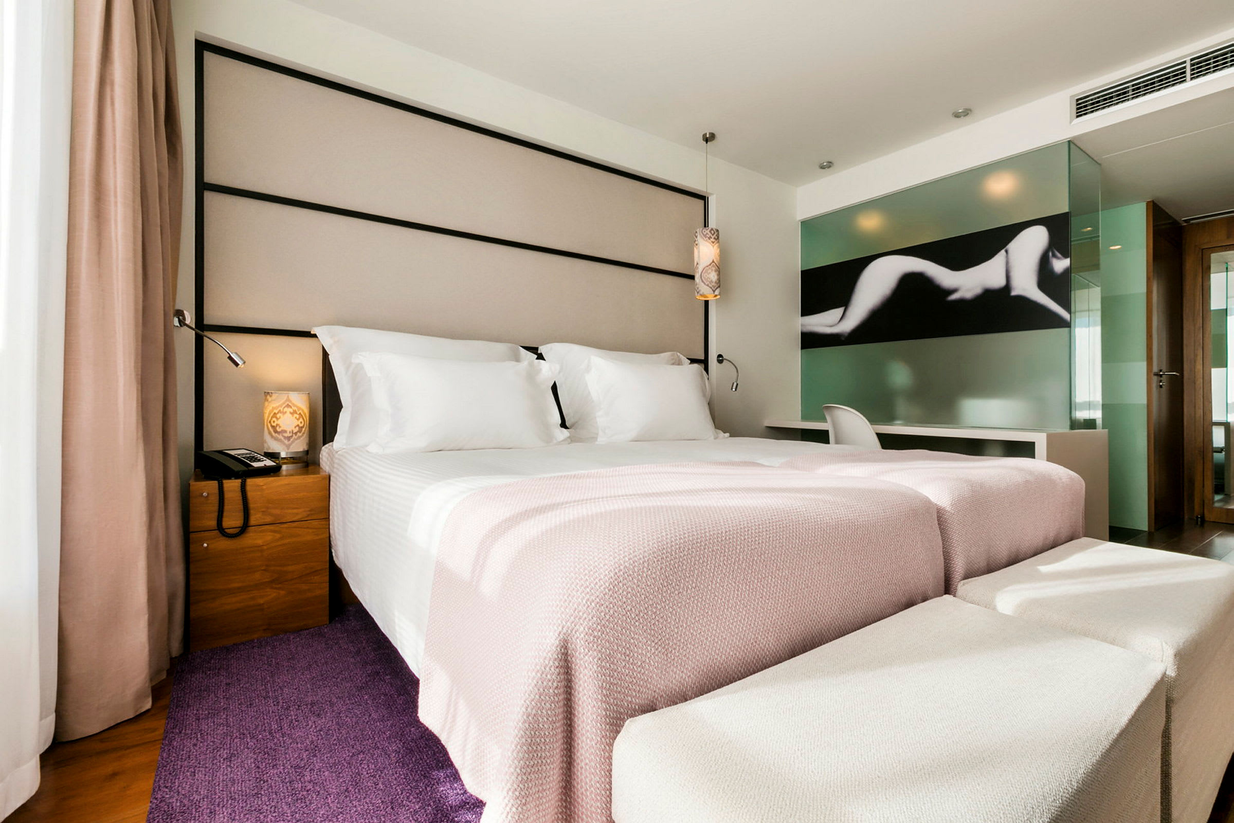 Moderni hotellihuone, jossa on leveä vuode, pedattu valkoisilla ja vaaleanpunasävyisillä vuodevaatteilla. Siinä on tuoli ja työpöytä lasiseinän vieressä, joka on koristeltu taidekuviolla. (Tekoälyn luoma teksti)