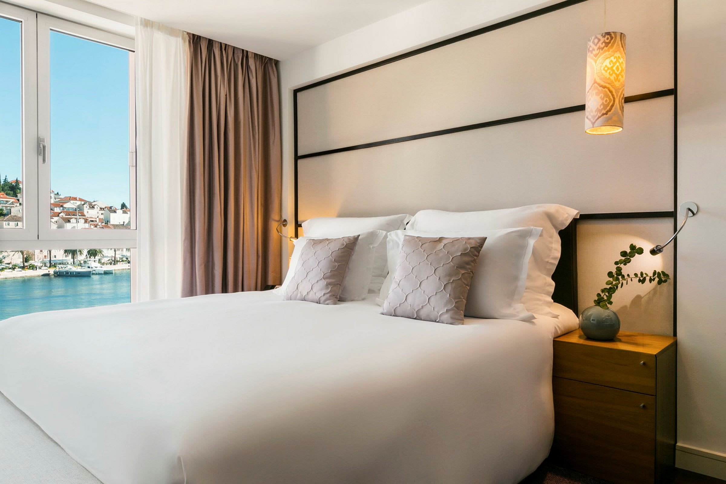 Hotellihuone, jossa on parisänky, koristetyynyt ja yöpöytä. Näkymä vedelle ja rakennuksiin ikkunan kautta. (Tekoälyn luoma teksti)