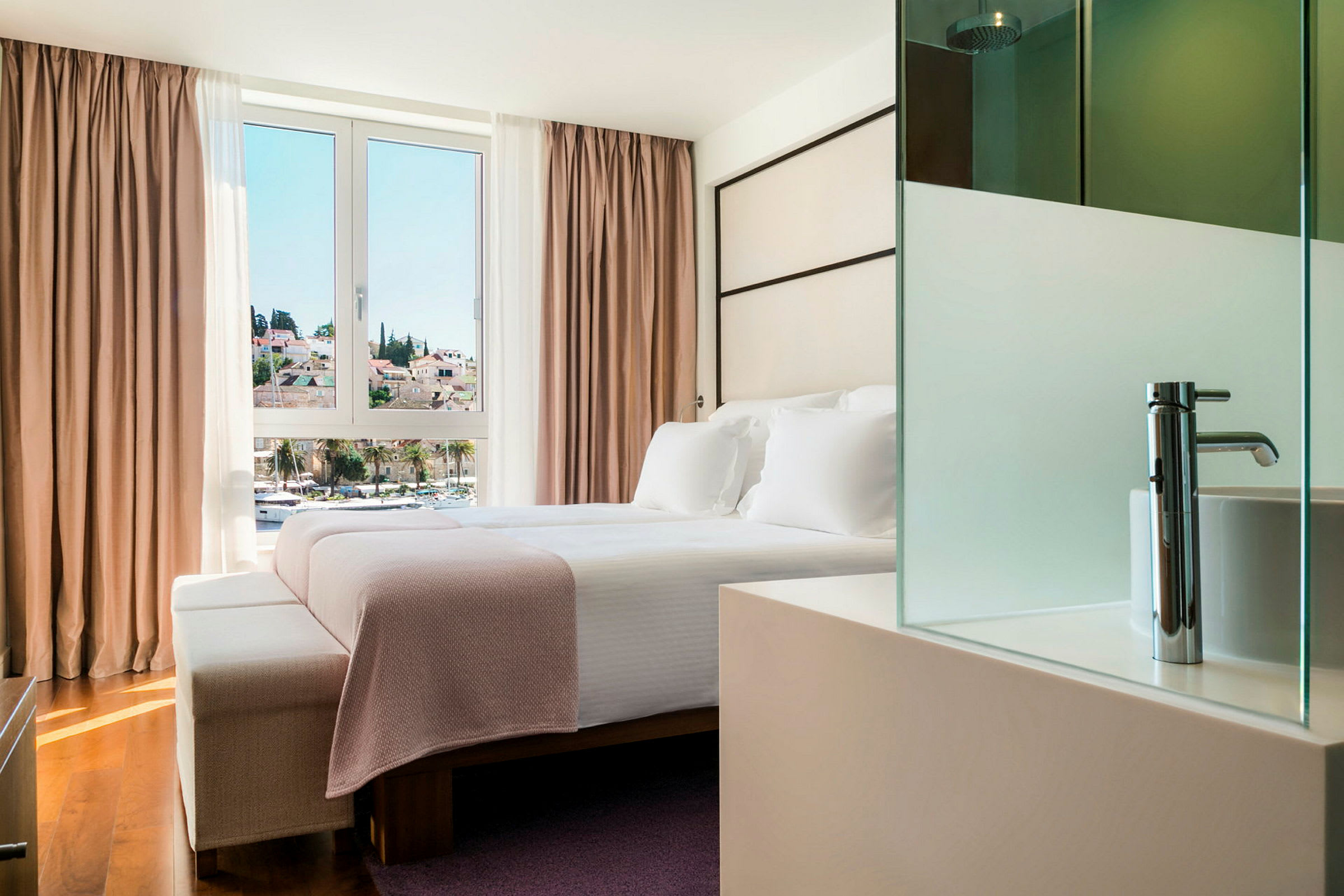 Hotellihuone, jossa on parisänky suuren ikkunan edessä, josta on näkymä kaupungille. Sänky on pedattu valkoisilla vuodevaatteilla ja vaaleanpunaisella huovalla. (Tekoälyn luoma teksti)