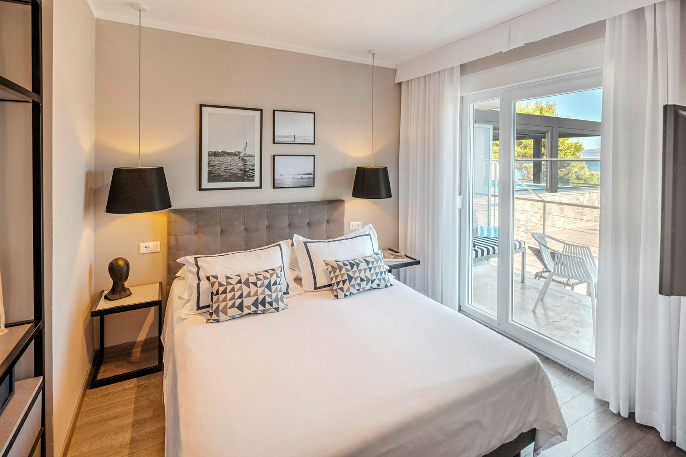 Moderni hotellihuone, jossa on parivuode, koristeltu tyynyillä ja taiteella seinällä. Lasiovet johtavat sisustetulle terassille. (Tekoälyn luoma teksti)