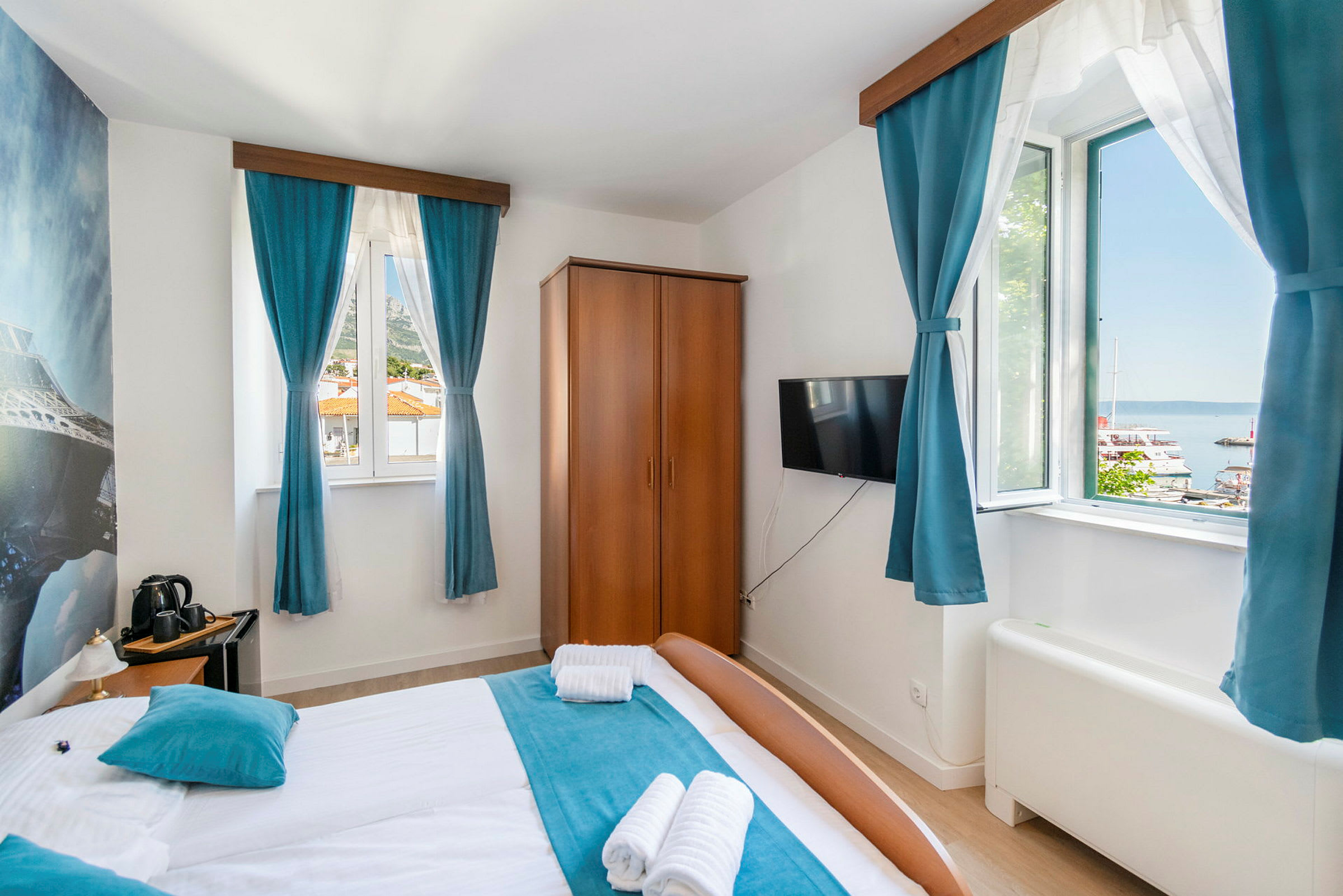 Hotellihuone, jossa on parivuode, siniset verhot ja näkymä satamaan ikkunan ulkopuolella. (Tekoälyn luoma teksti)