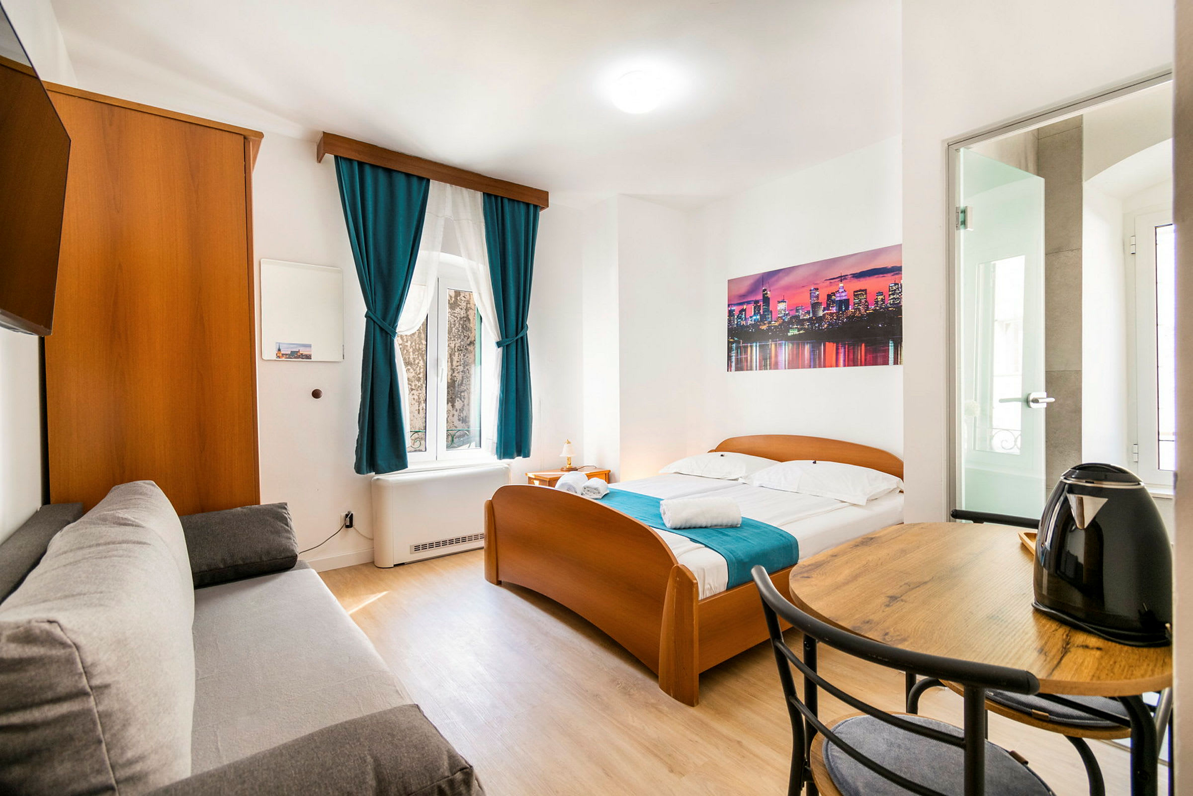Kuva valoisasta hotellihuoneesta, jossa on sänky, sohva, pöytä ja vedenkeitin. (Tekoälyn luoma teksti)