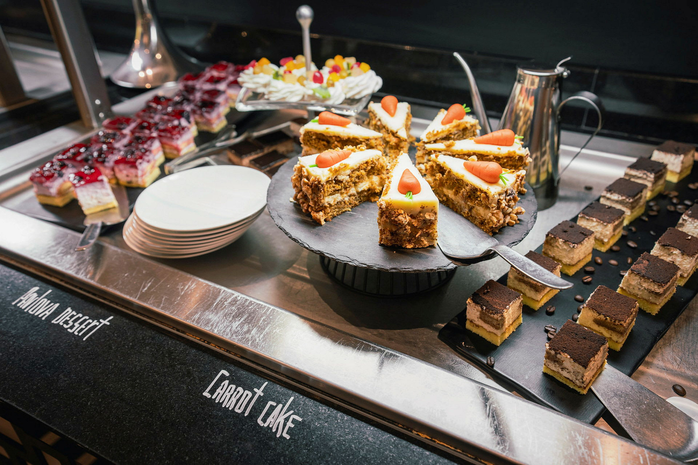 Kuvassa näkyy buffet, jossa on erilaisia jälkiruokia, mukaan lukien porkkanakakku, pienet vanilja- ja suklaaleivonnaiset sekä värikkäitä hedelmäkarkkeja. (Tekoälyn luoma teksti)