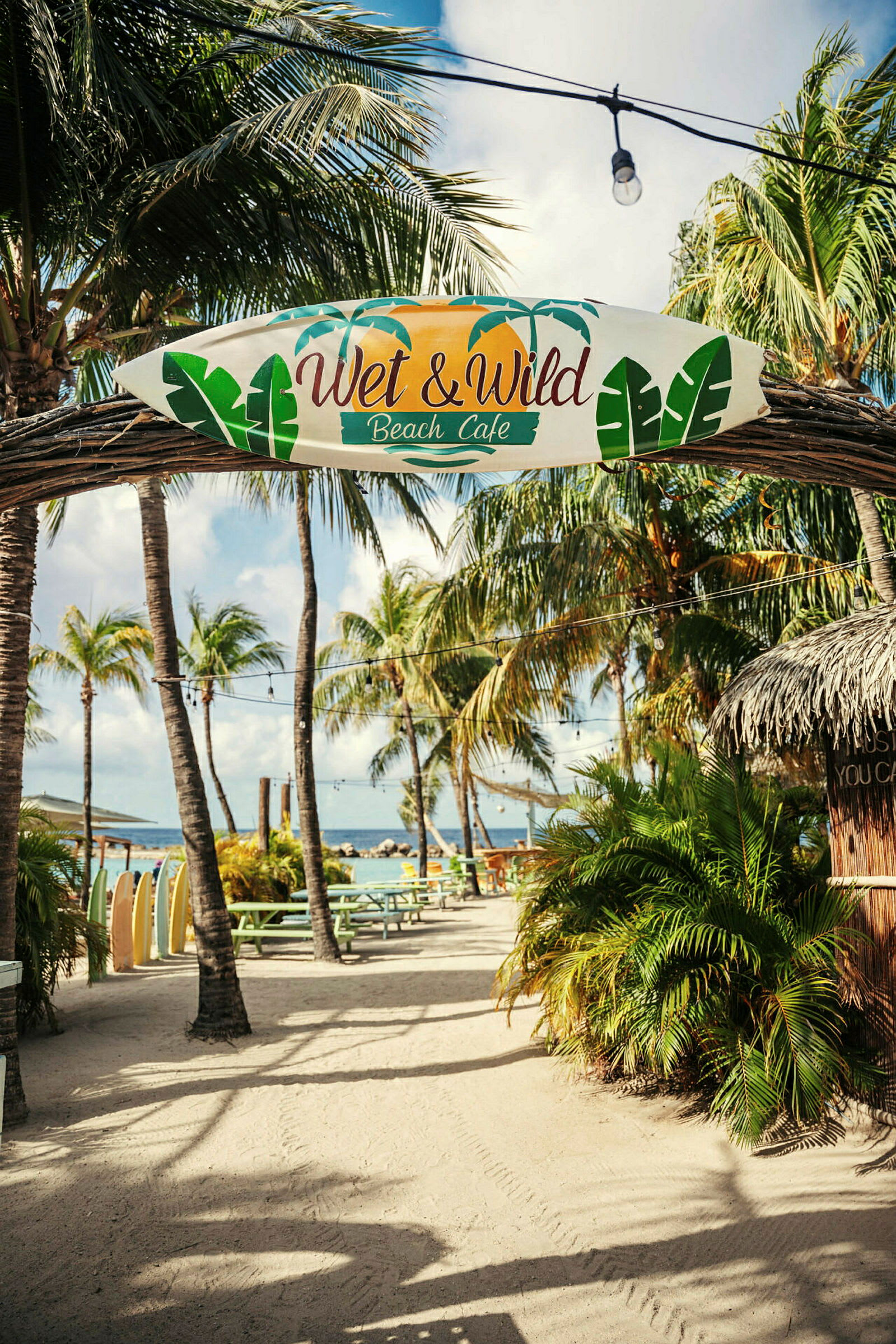 "Wet & Wild Beach Café" -kyltti riippuu kaarella ulkoilmassa, taustalla palmuja, pöytiä ja meri. (Tekoälyn luoma teksti)