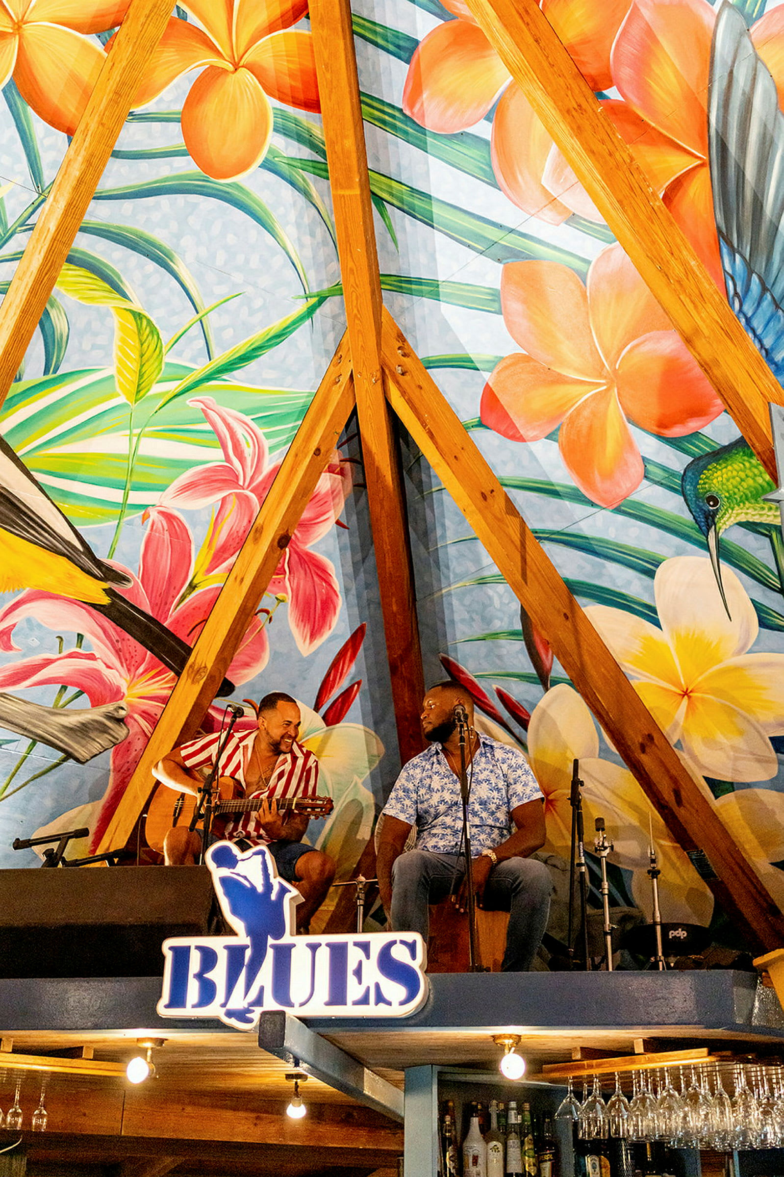 Värikäs seinämaalaus, jossa on kukkia ja lintuja, koristaa seinää puurakenteessa lavan yläpuolella, jossa on soittimia. (Tekoälyn luoma teksti)