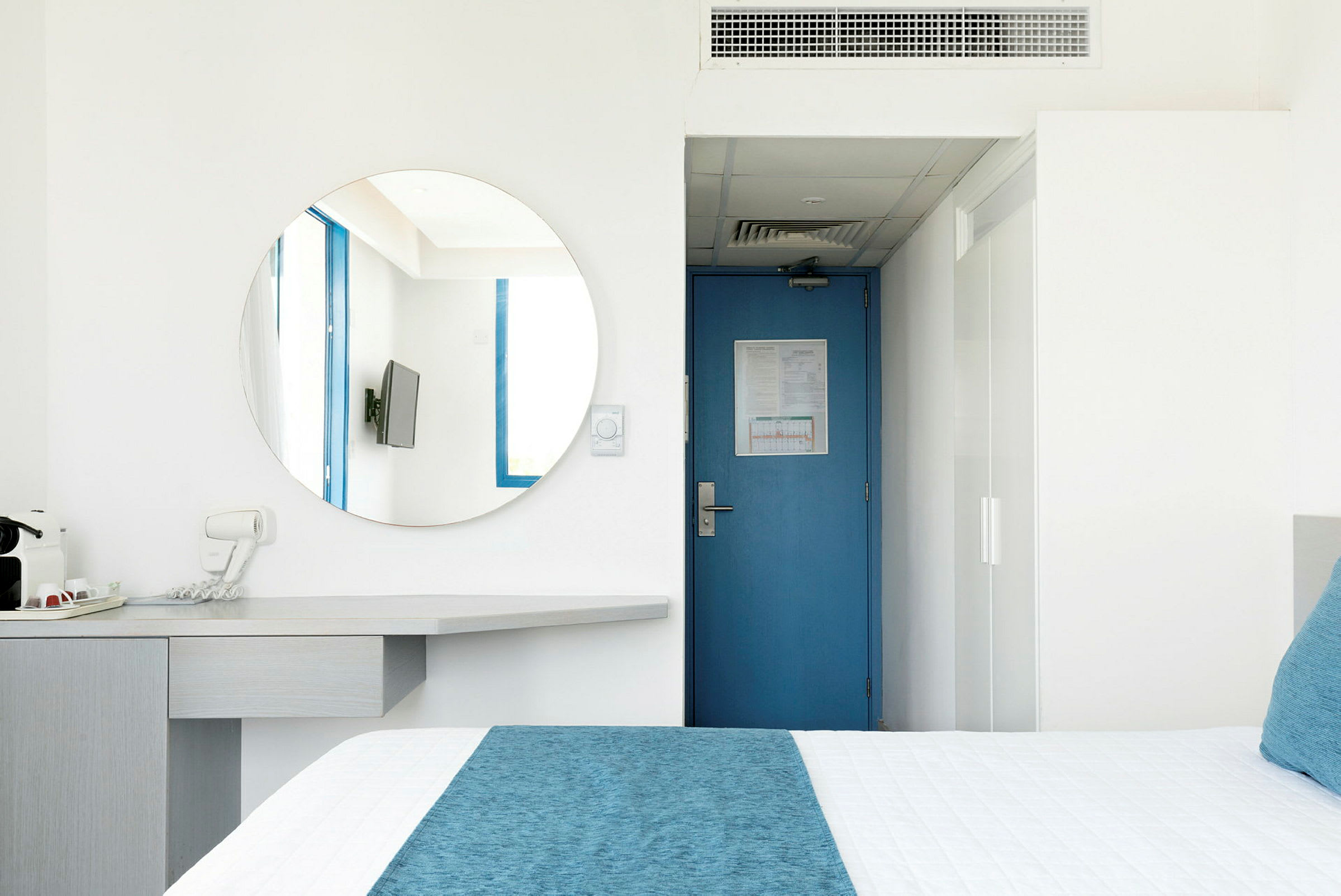 Hotellihuone, jossa sininen ovi, pyöreä peili, televisio ja sinisiä yksityiskohtia sisältävä sänky. (Tekoälyn luoma teksti)