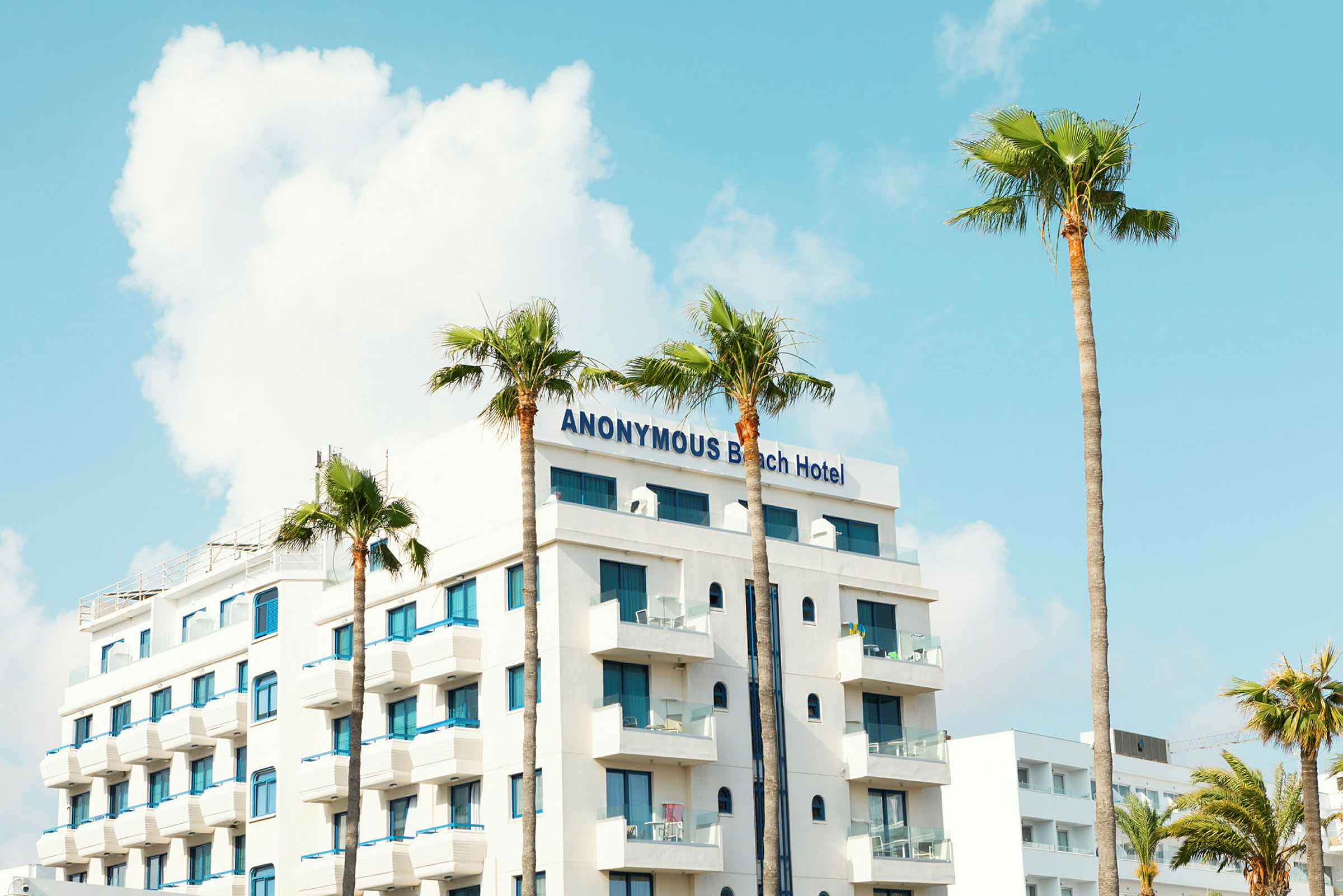 Anonymous Beach Hotel -niminen hotelli, jossa on valkoinen julkisivu ja siniset ikkunat, ympärillä palmuja. (Tekoälyn luoma teksti)