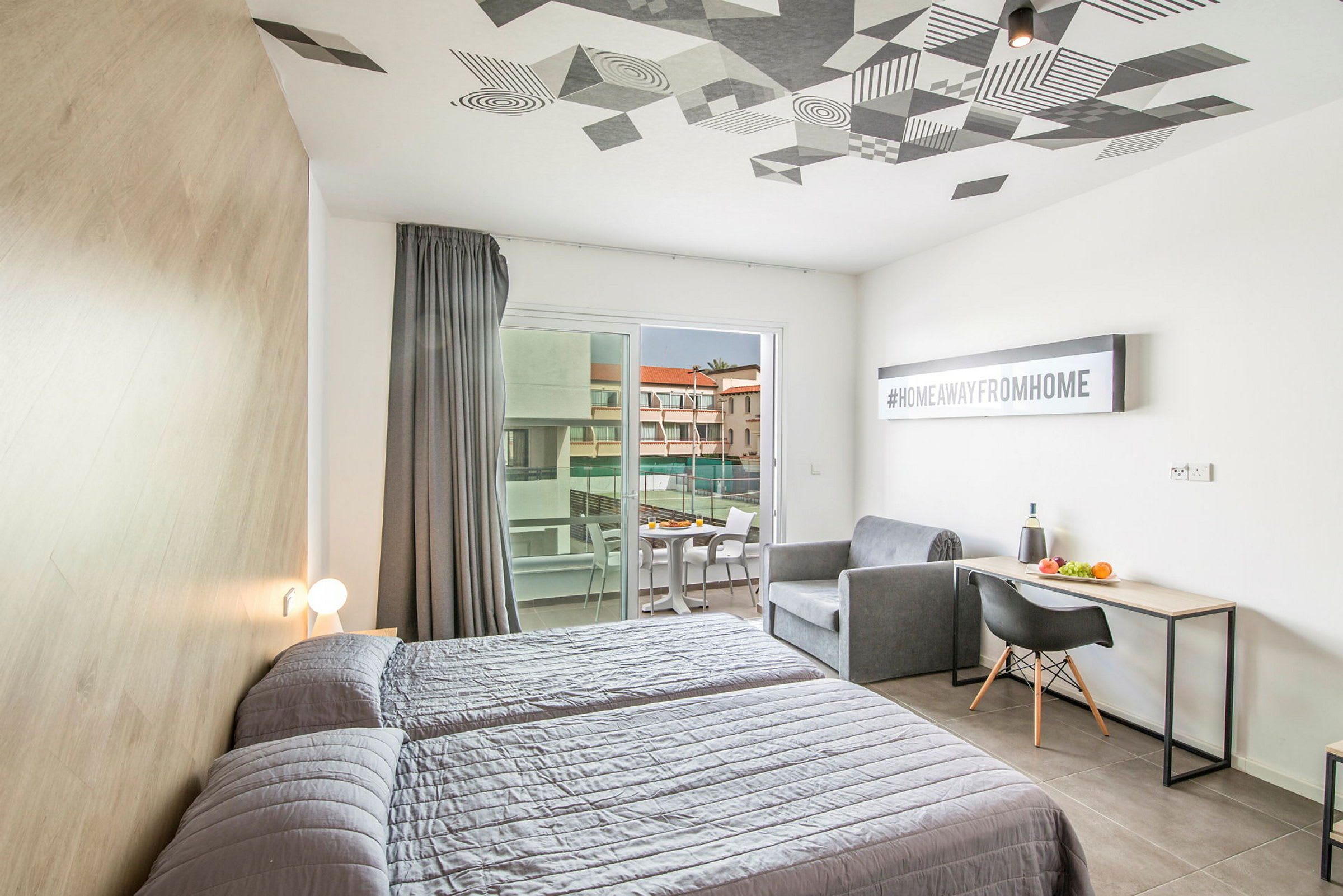 Hotellihuone, jossa on kaksi sänkyä, sohva, kirjoituspöytä ja parveke, jolta avautuu näkymä rakennukseen. (Tekoälyn luoma teksti)
