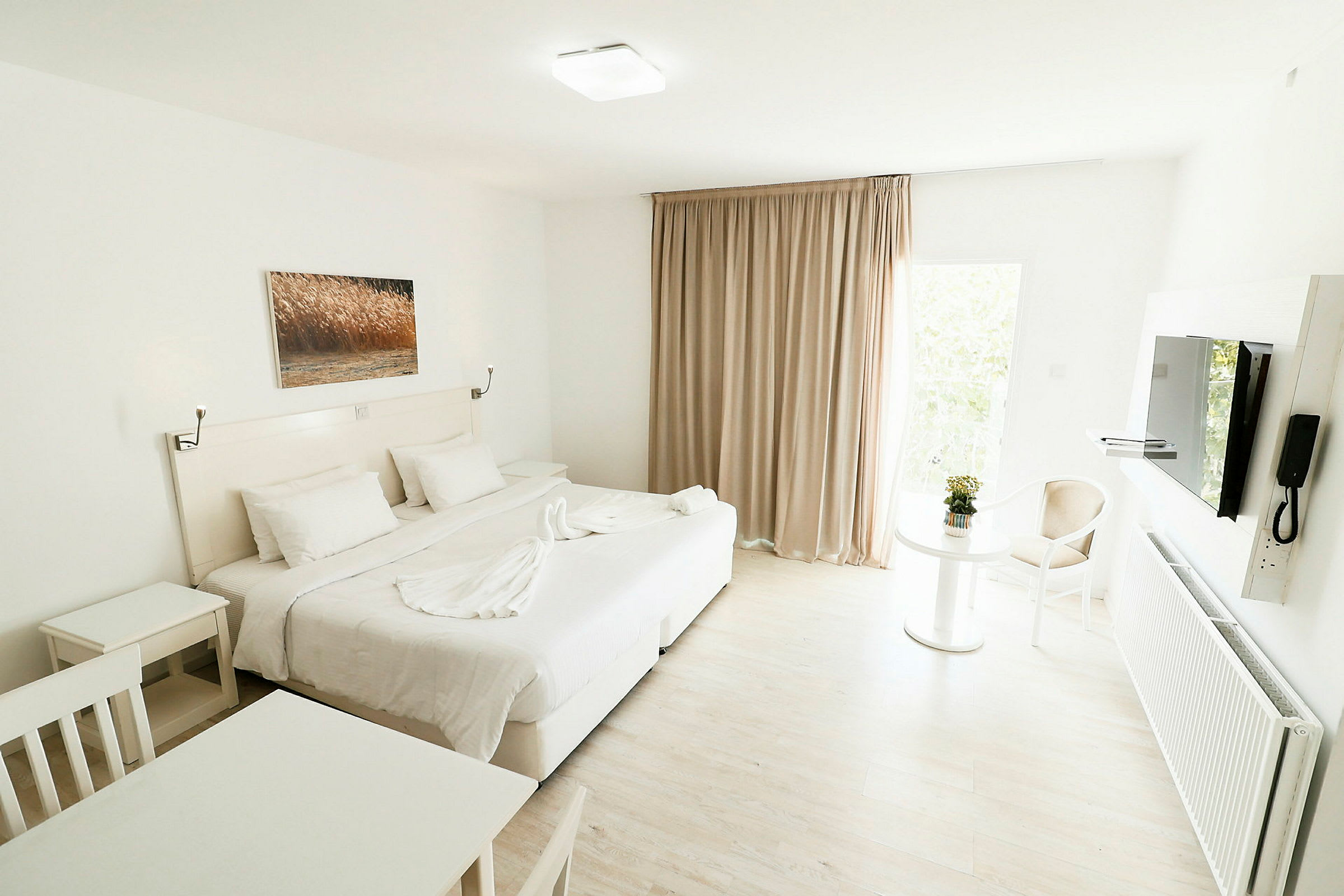 Valoisa ja moderni hotellihuone, jossa on suuri sänky, valkoiset huonekalut ja taulu seinällä. Pöytä, jossa kukka ja tuoli, näkyy ikkunan ääressä beigejen verhojen kanssa. (Tekoälyn luoma teksti)
